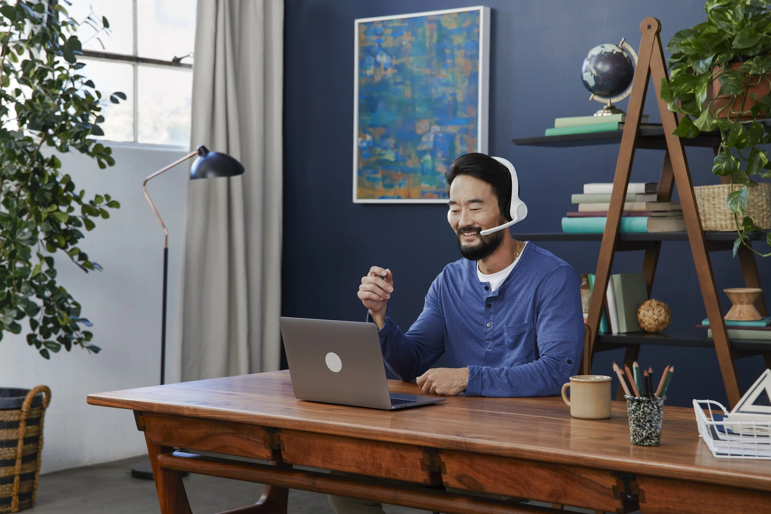 A man sitting at a wooden desk with a laptop, wearing a white headset, smiling and looking at the screen. The background includes a dark blue wall with a colorful abstract painting, a black shelf with books and a globe, a potted plant, and a large wi