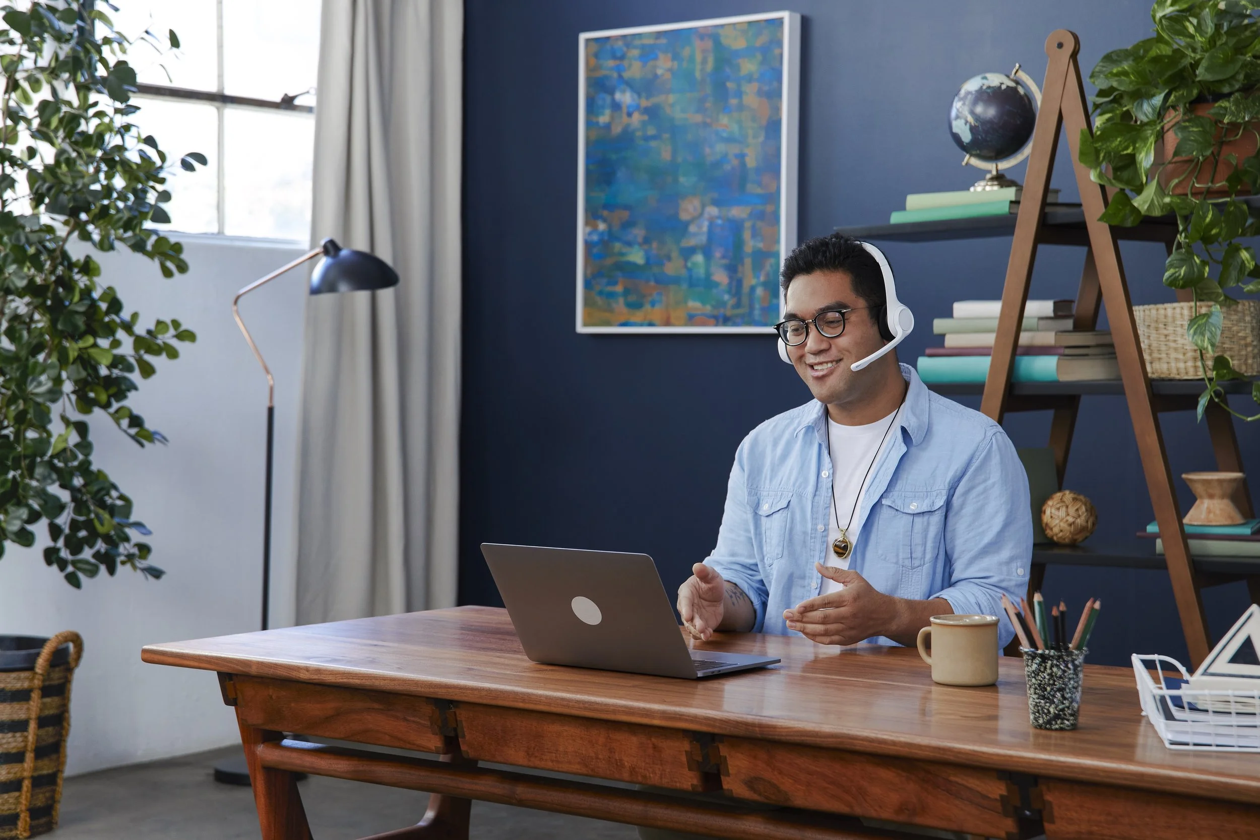 A man with glasses and headphones sitting at a wooden desk, smiling and looking at a laptop. The room has a blue wall with a colorful abstract painting, a bookshelf with books and plants, a window with white curtains, and a black desk lamp.