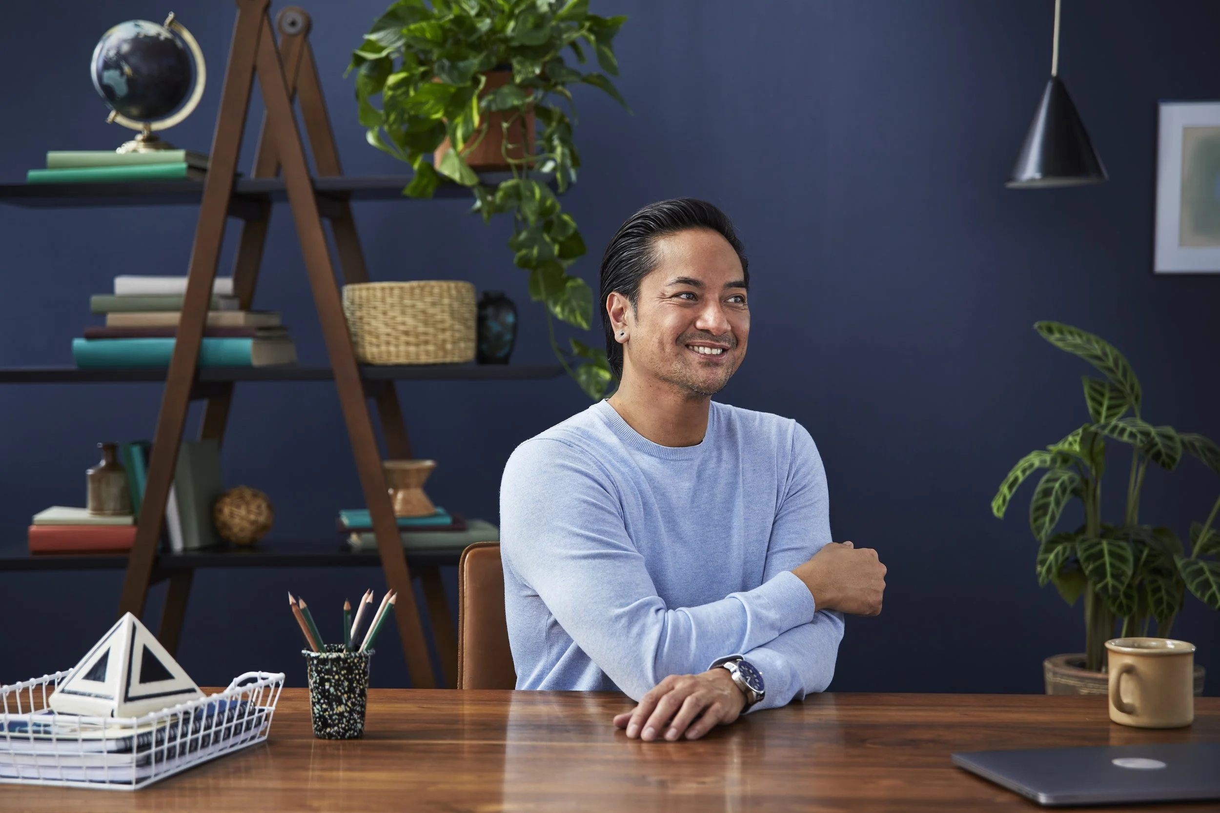 A man with dark hair wearing a light blue long-sleeved shirt, sitting at a wooden table with his arms crossed and smiling, in a room with the blue wall, shelves with books, a globe, plants, and various decorative items.