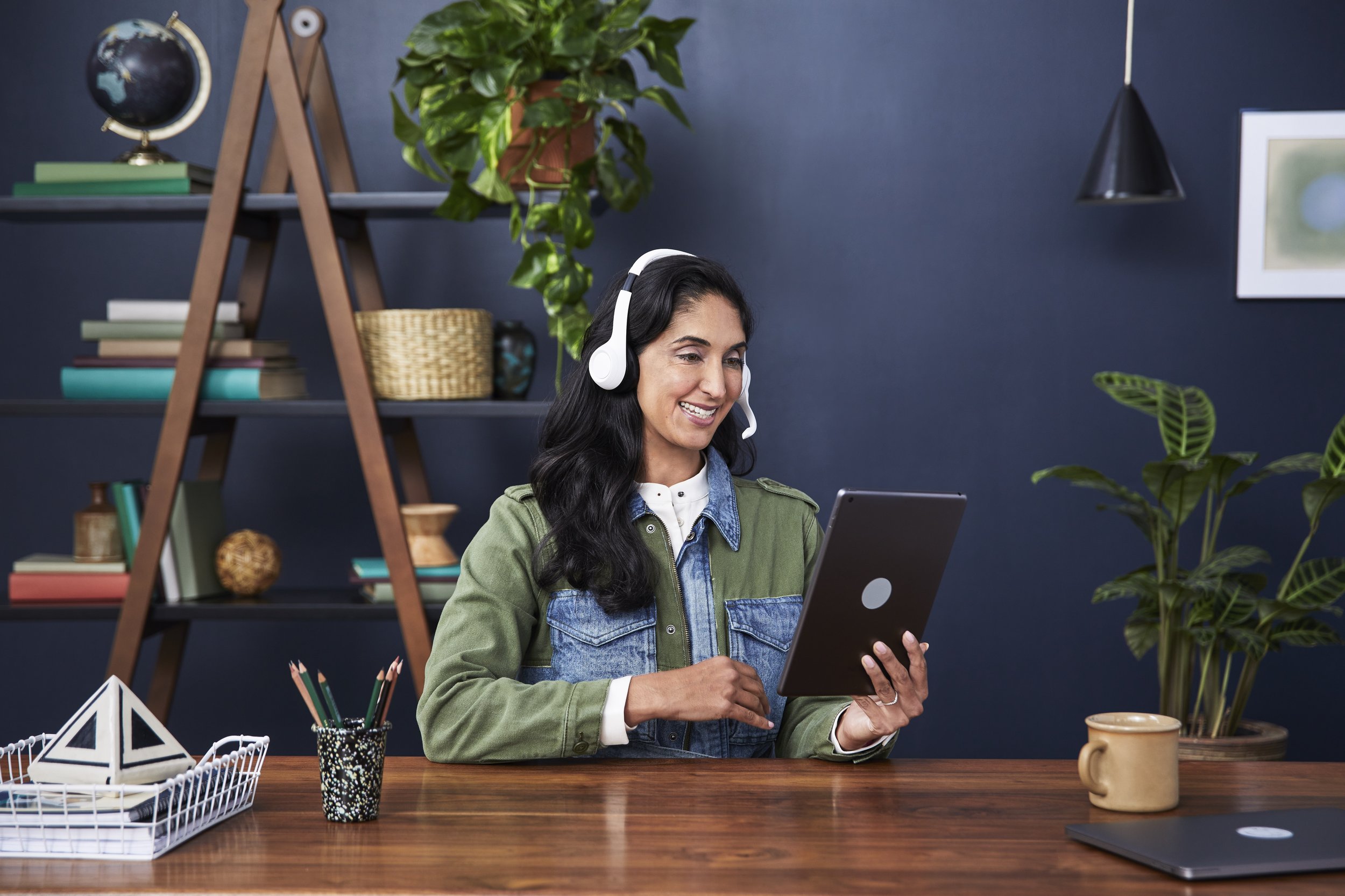 A woman with dark hair and light skin wearing headphones, a denim jacket, and a green shirt, smiling and looking at a tablet in a cozy, decorated home office.