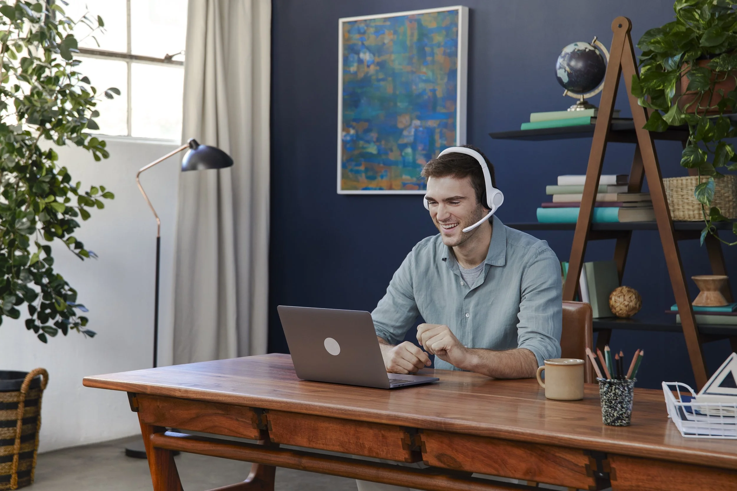A young man wearing a white headset smiling while working on a laptop at a wooden desk in a home office, with plants, books, and artwork in the background.