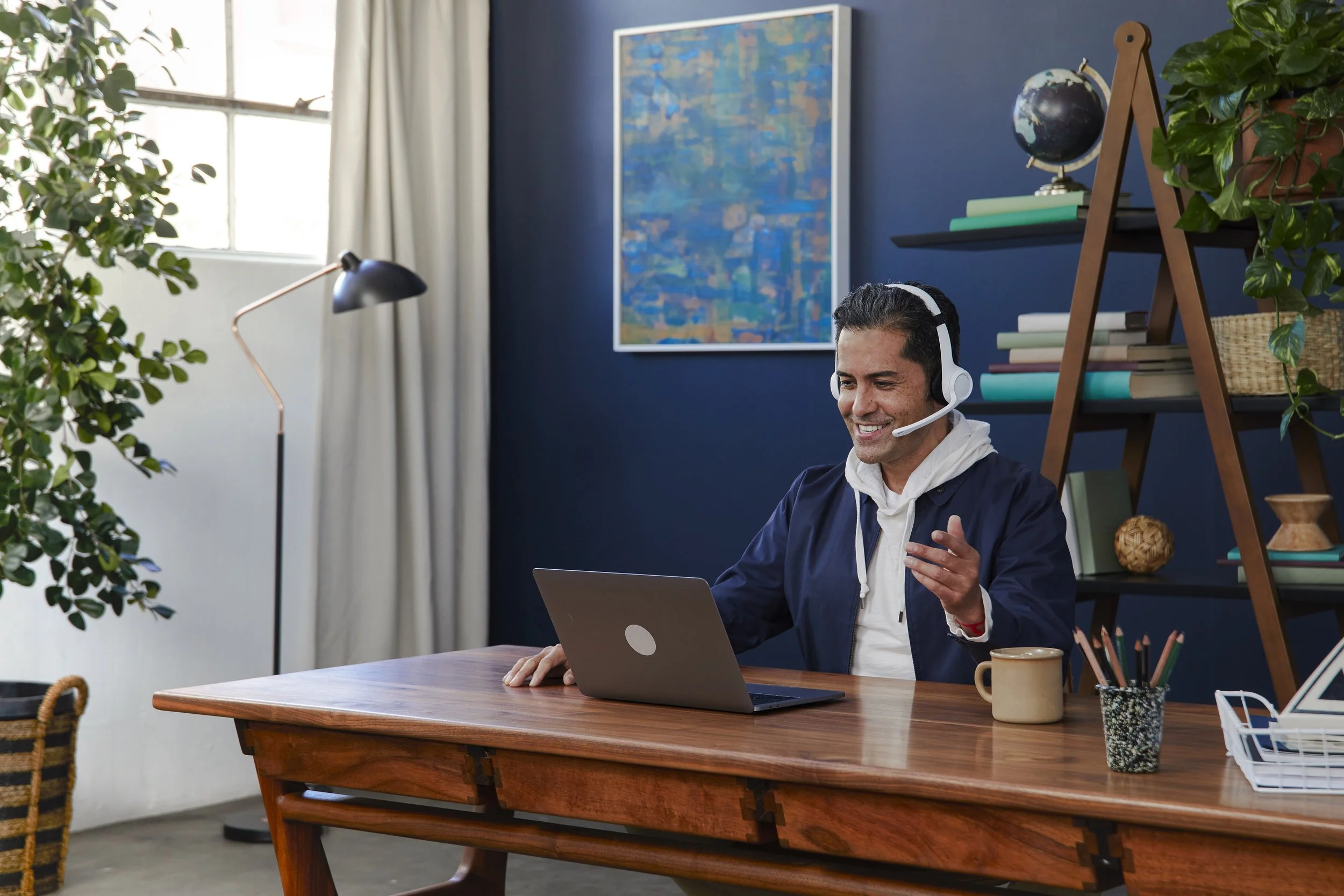 A man sitting at a wooden desk wearing a headset, smiling, with a laptop in front of him in a home office with plants, books, and colorful artwork on the wall.