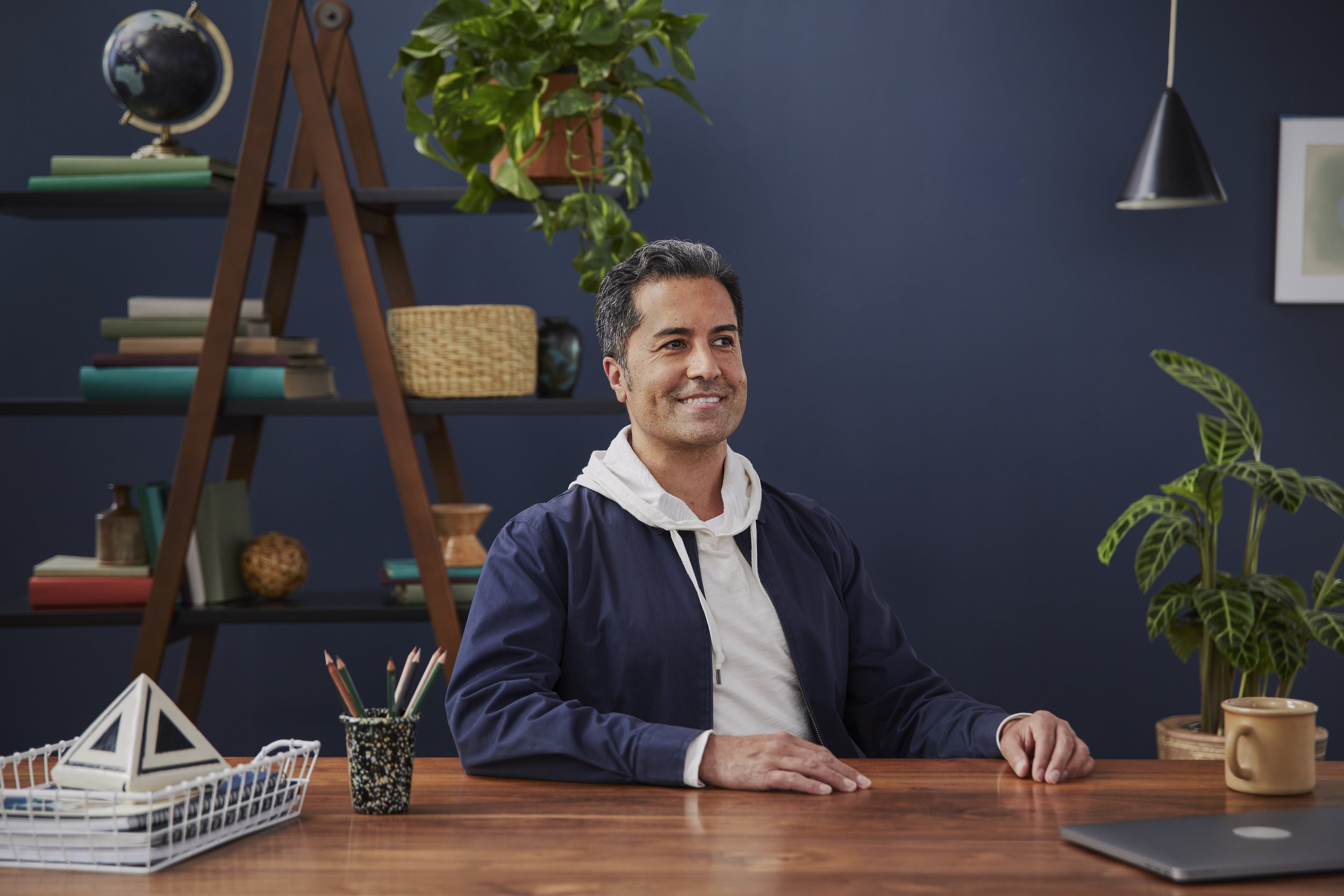 A man is sitting at a wooden desk in a room decorated with blue walls, surrounded by green plants, books, and decorative objects. He is smiling and wearing a dark jacket over a white hoodie.