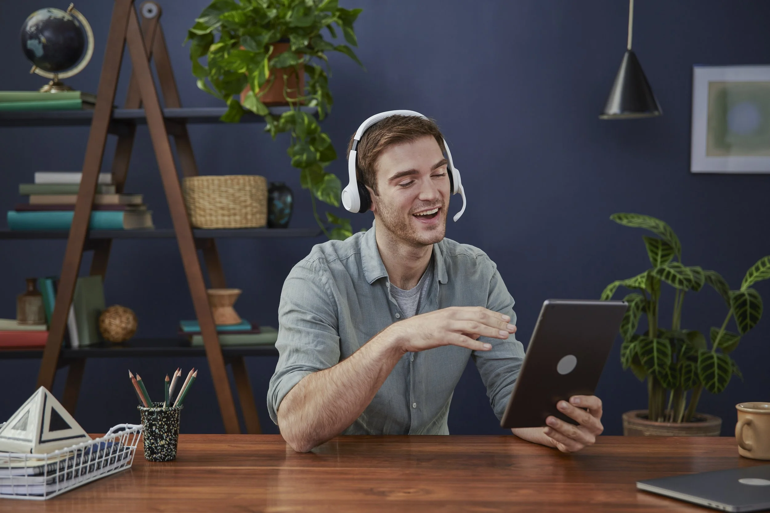 Young man smiling, wearing a headset, sitting at a desk with a tablet in his hand, in a home office decorated with plants, books, and office supplies.