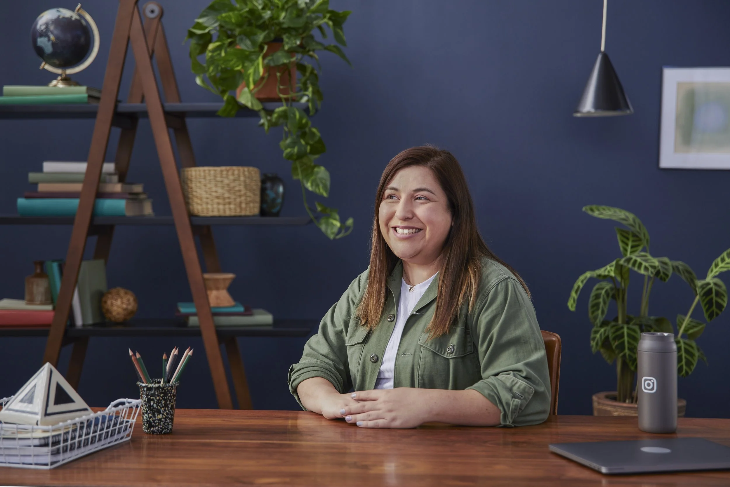 A woman with long brown hair smiling, sitting at a wooden desk with plants and office supplies, in a room with a dark blue wall and a bookshelf.