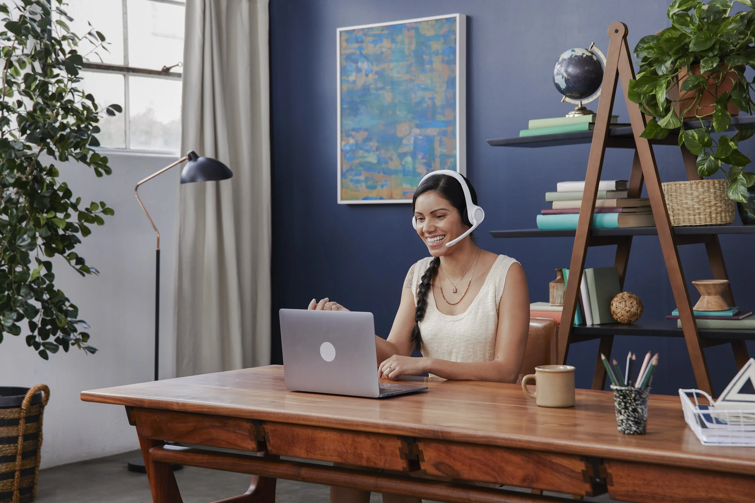 A woman smiling and wearing a headset while working on a laptop at a wooden desk in a home office with a blue wall, a large window, a bookshelf with books and plants, and a framed abstract painting.