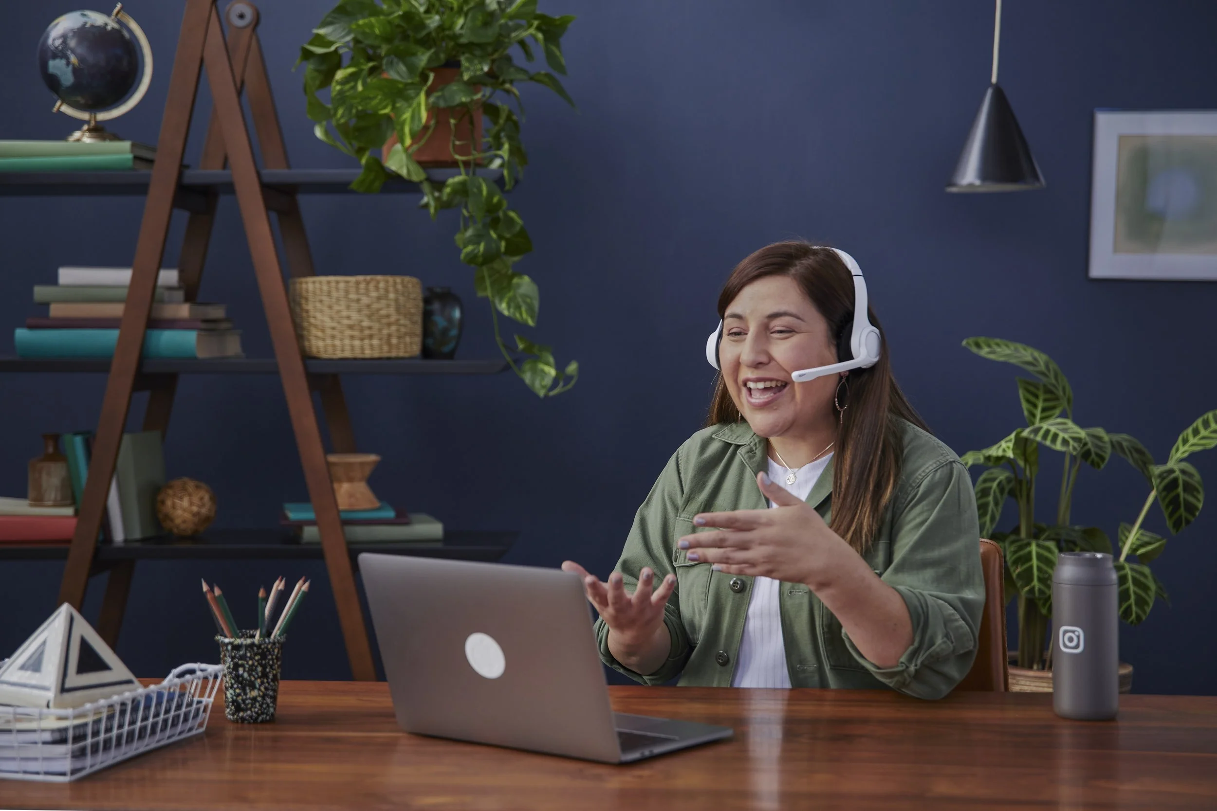 A woman with brown hair wearing a green jacket and a headset sitting at a desk, smiling and gesturing with her hands, with a laptop and various office supplies on the desk, in a room decorated with plants and shelves.