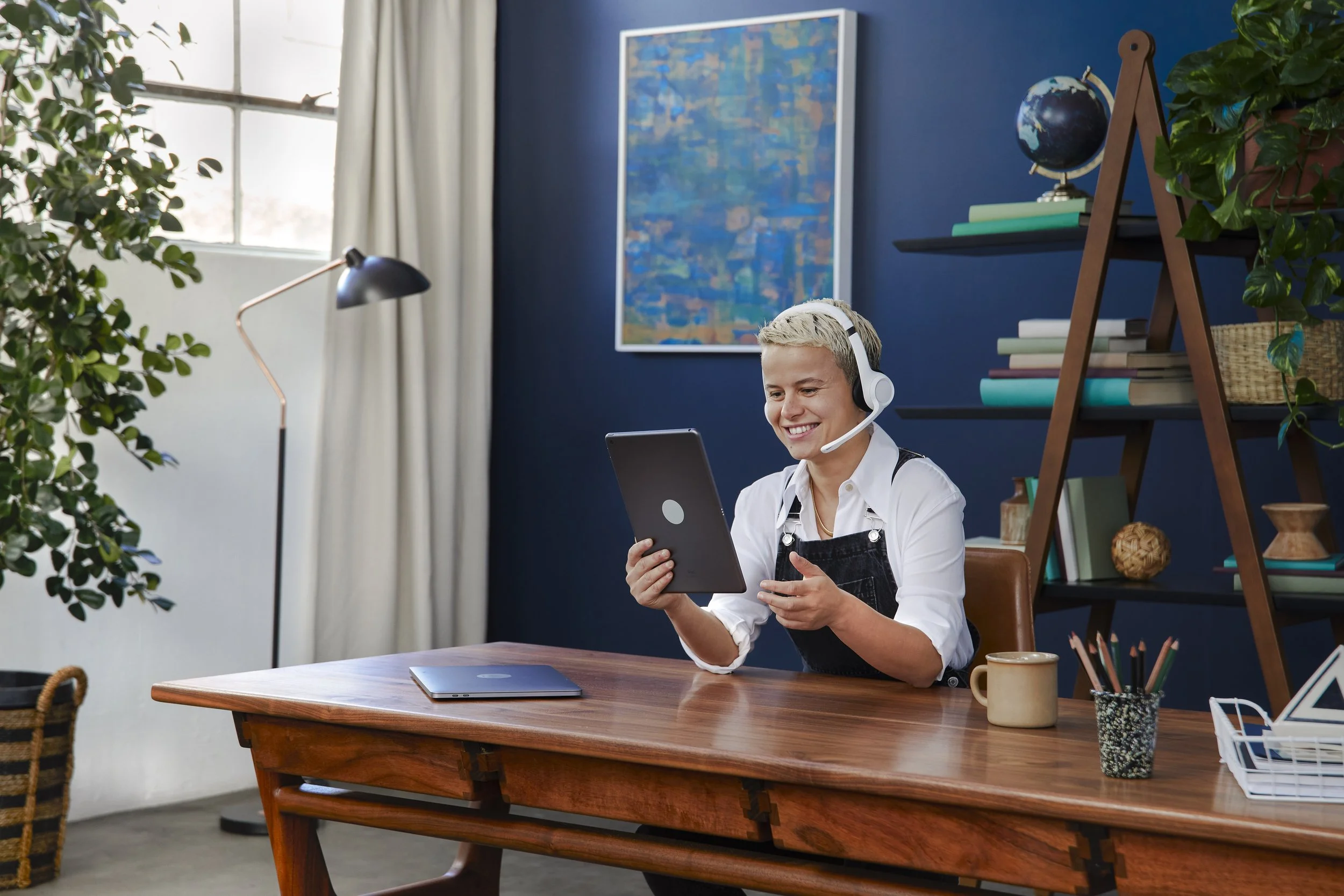A woman wearing a white shirt, black apron, and headset is smiling while looking at a tablet in a cozy, well-lit room with blue walls, a large window, and a decorated bookshelf. There are plants, a laptop, a mug, and various office supplies on the wo