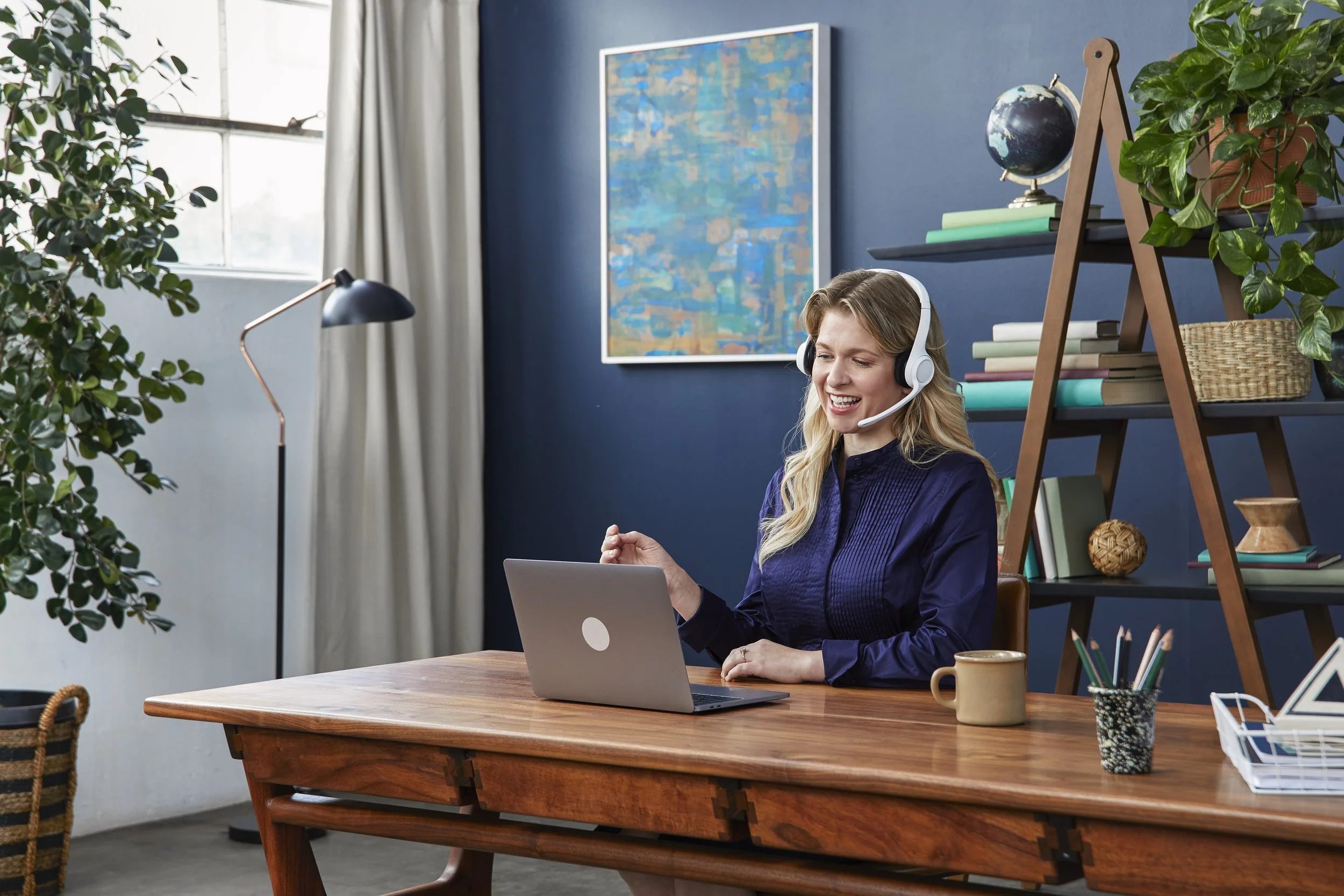 A woman wearing a headset sitting at a wooden desk with a laptop, smiling and looking at the screen, in a room with plants, books, and a large abstract painting on a blue wall.