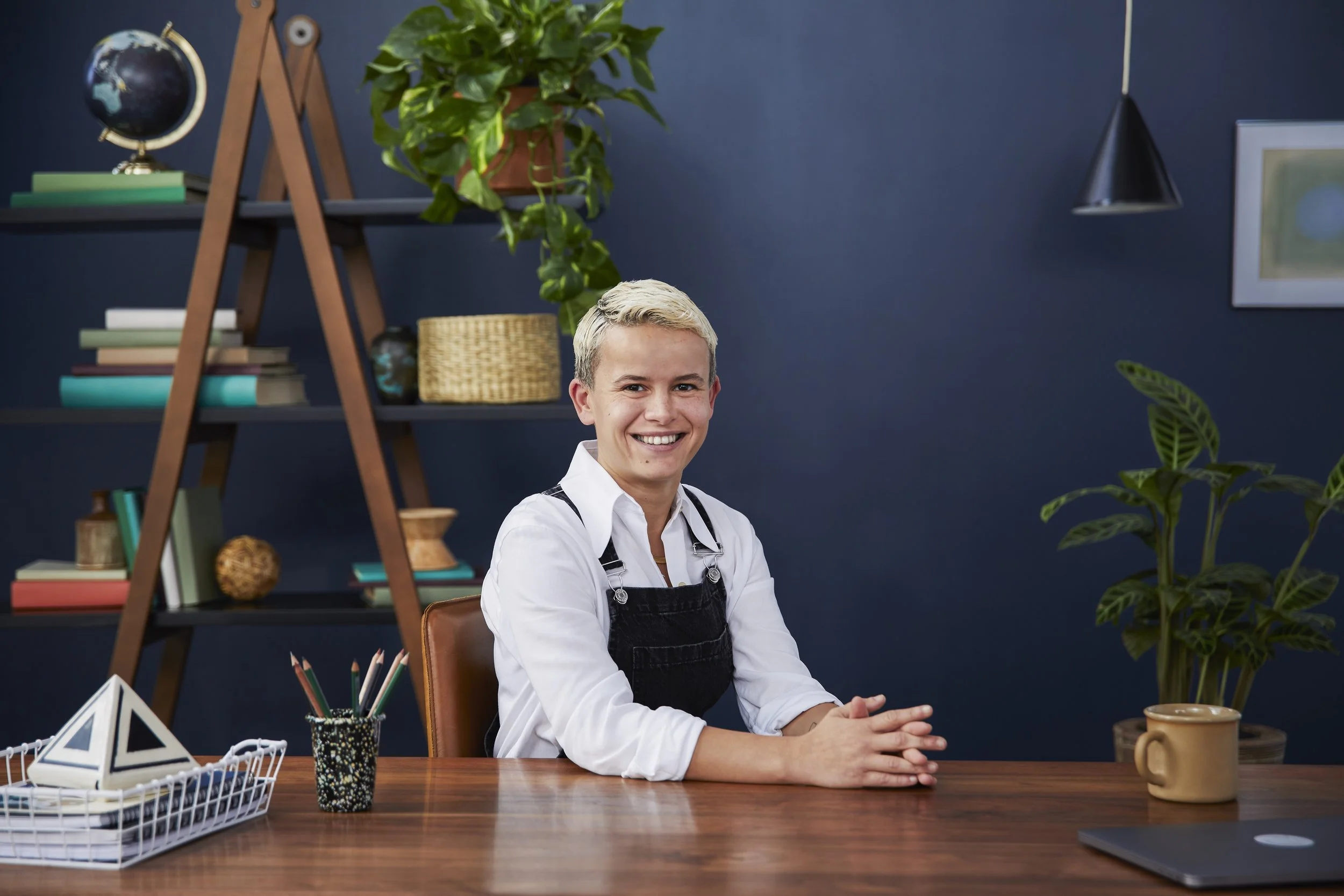 A young woman with blonde hair smiling and sitting at a wooden desk in a modern office space. She is wearing a white shirt and black apron. In the background, there are shelves with books, a globe, and potted plants. The desk has a basket of geometri