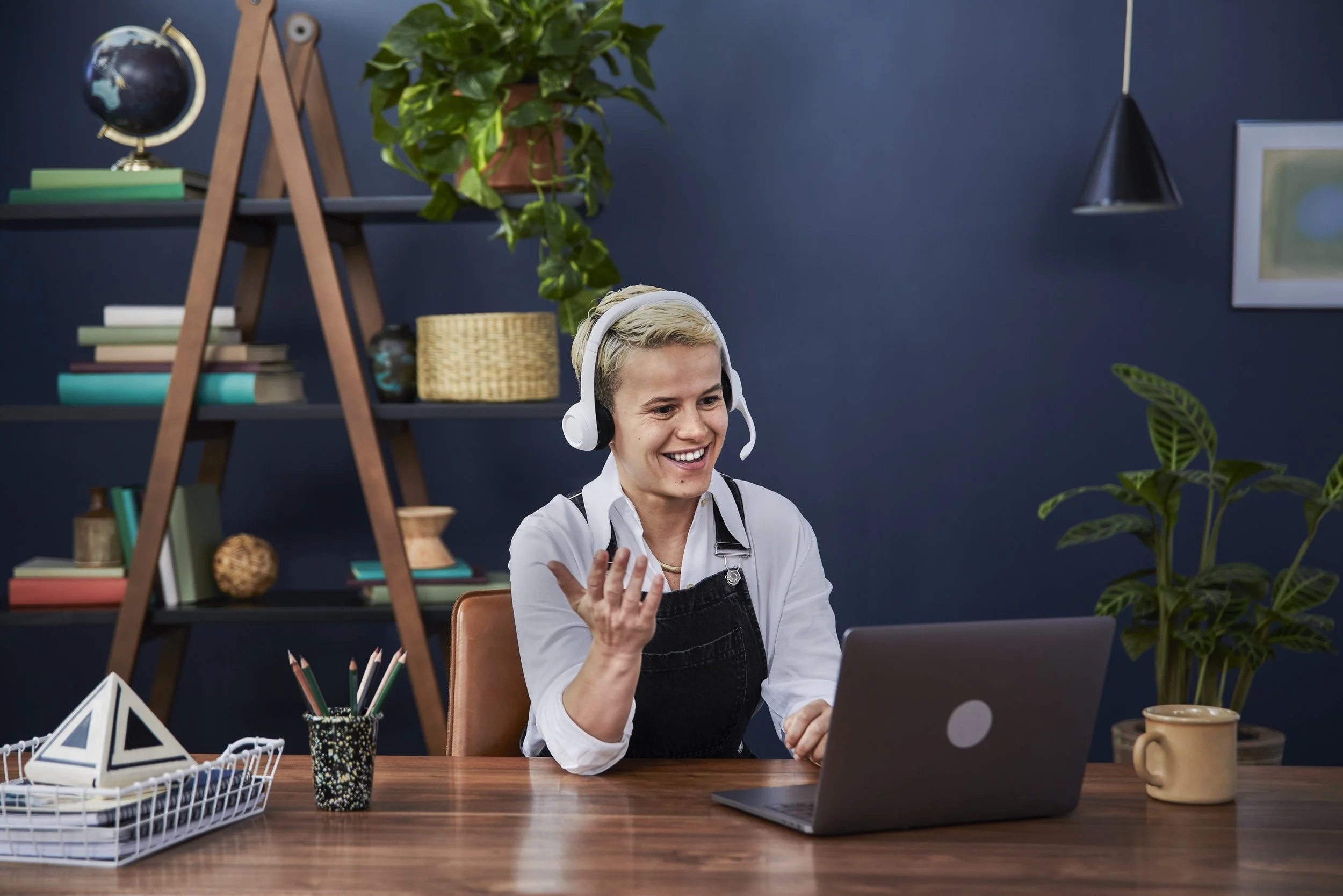 A woman with short blonde hair, wearing white shirt and black apron, smiling and talking while wearing headphones. She is seated at a wooden desk with a laptop and various office supplies. Behind her is a dark blue wall with a bookshelf, plants, and 