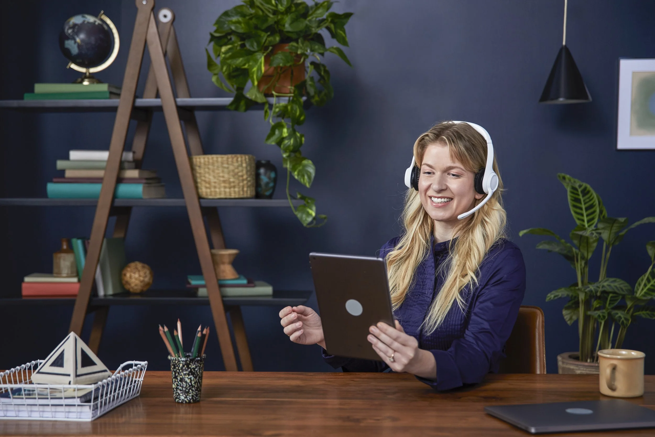 A smiling woman with blonde hair wearing a headset and holding a tablet, sitting at a wooden desk with various office supplies, plants, and decorative items in a home office with navy blue walls.