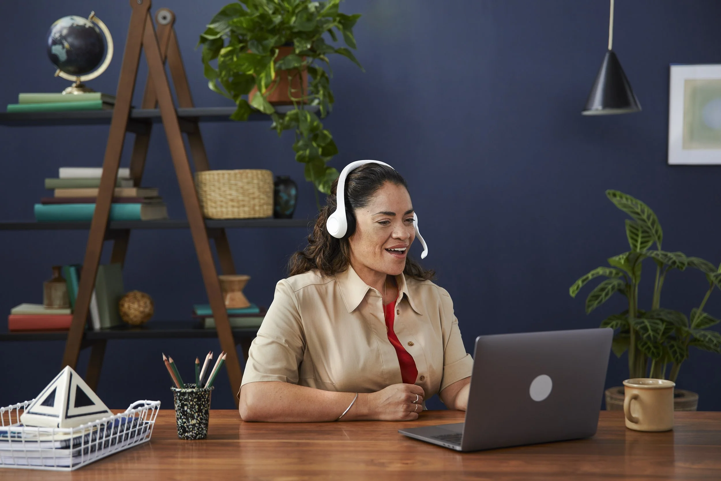 A woman with dark hair wearing a beige shirt and red top, smiling and looking at her laptop while wearing a white headset, seated at a wooden desk with a potted plant, a geometric paper decoration, a container of pencils, and a document holder.