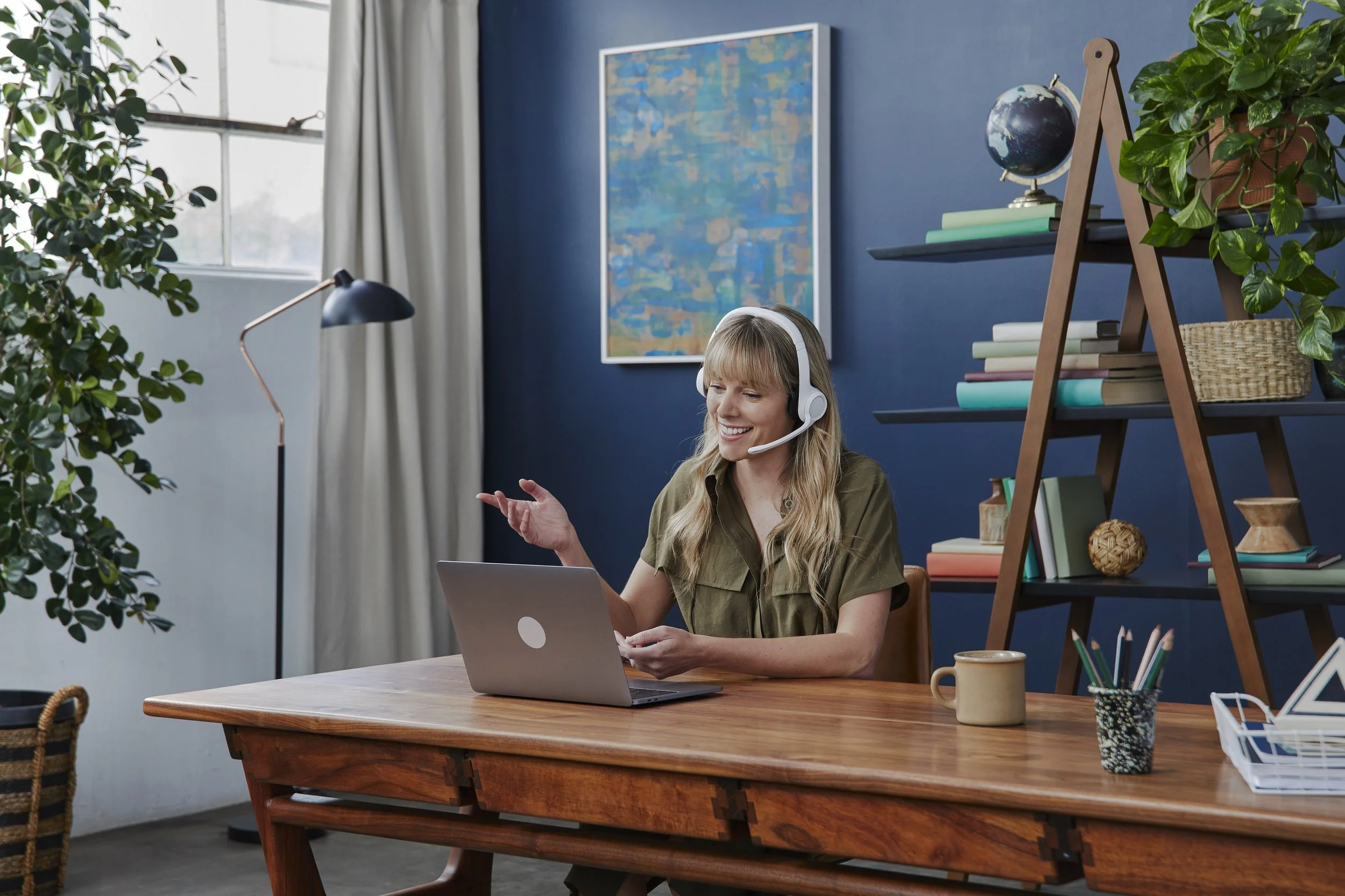 A woman with long blonde hair and wearing a headset is sitting at a wooden desk, smiling and looking at her phone, with a laptop open in front of her. The room has a blue wall, a window with curtains, a bookshelf with books and decorative items, and 