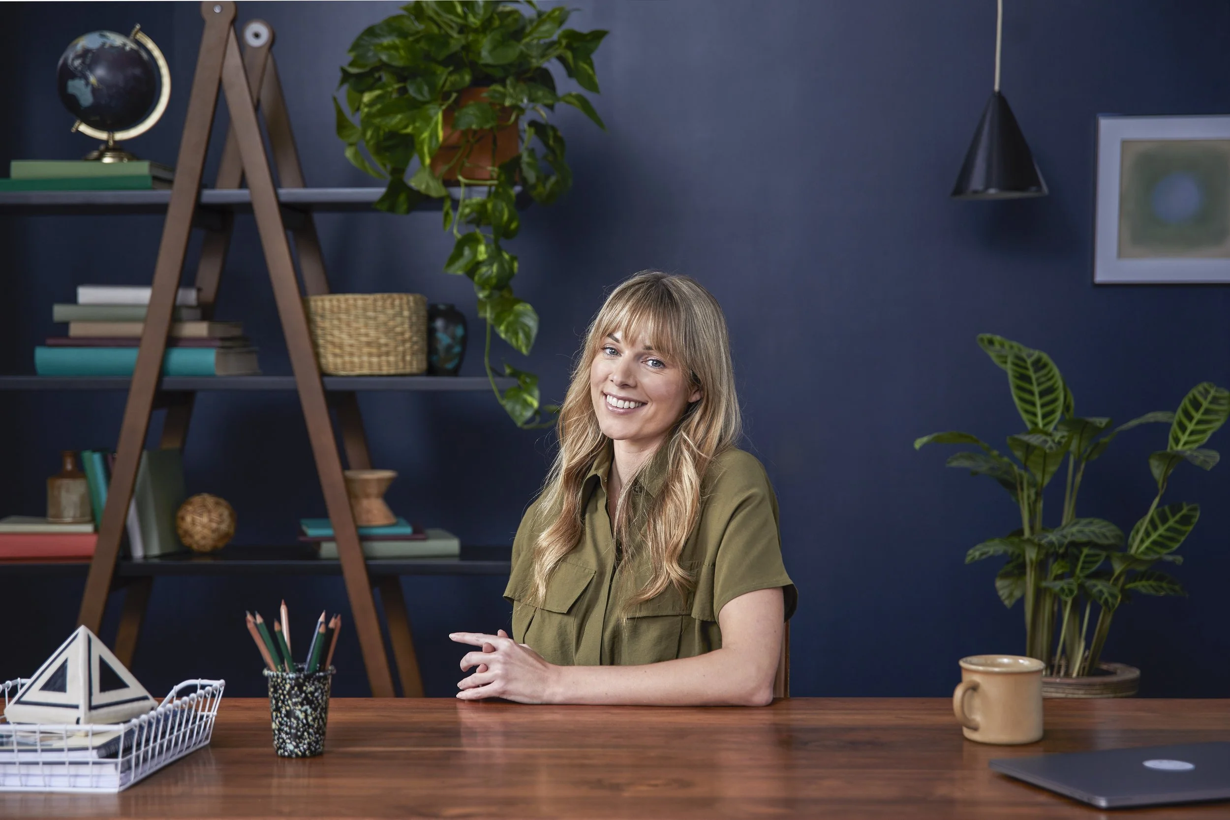 Young woman with blonde hair smiling and sitting at a desk with plants, books, and office supplies in a home office environment.