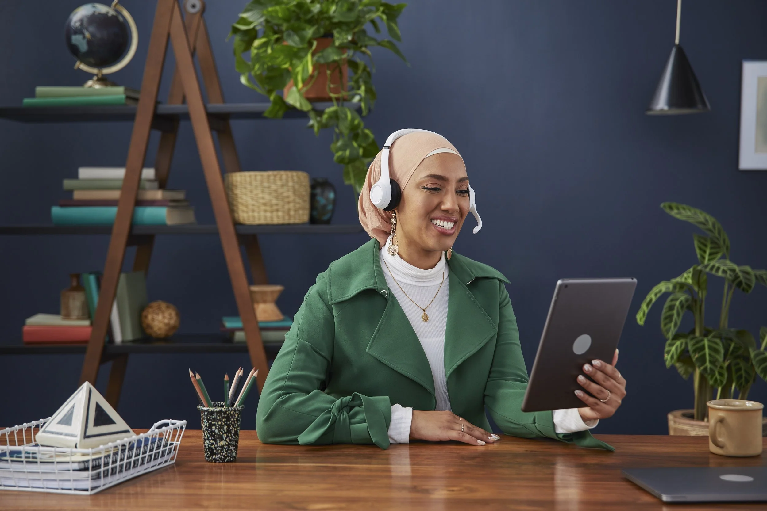 A woman wearing a light pink headscarf, white turtleneck, and green blazer sits at a wooden desk, smiling while holding a tablet. She has earrings and a necklace. On the desk are pencils in a cup, a white geometric-shaped decor item, a wire basket, a