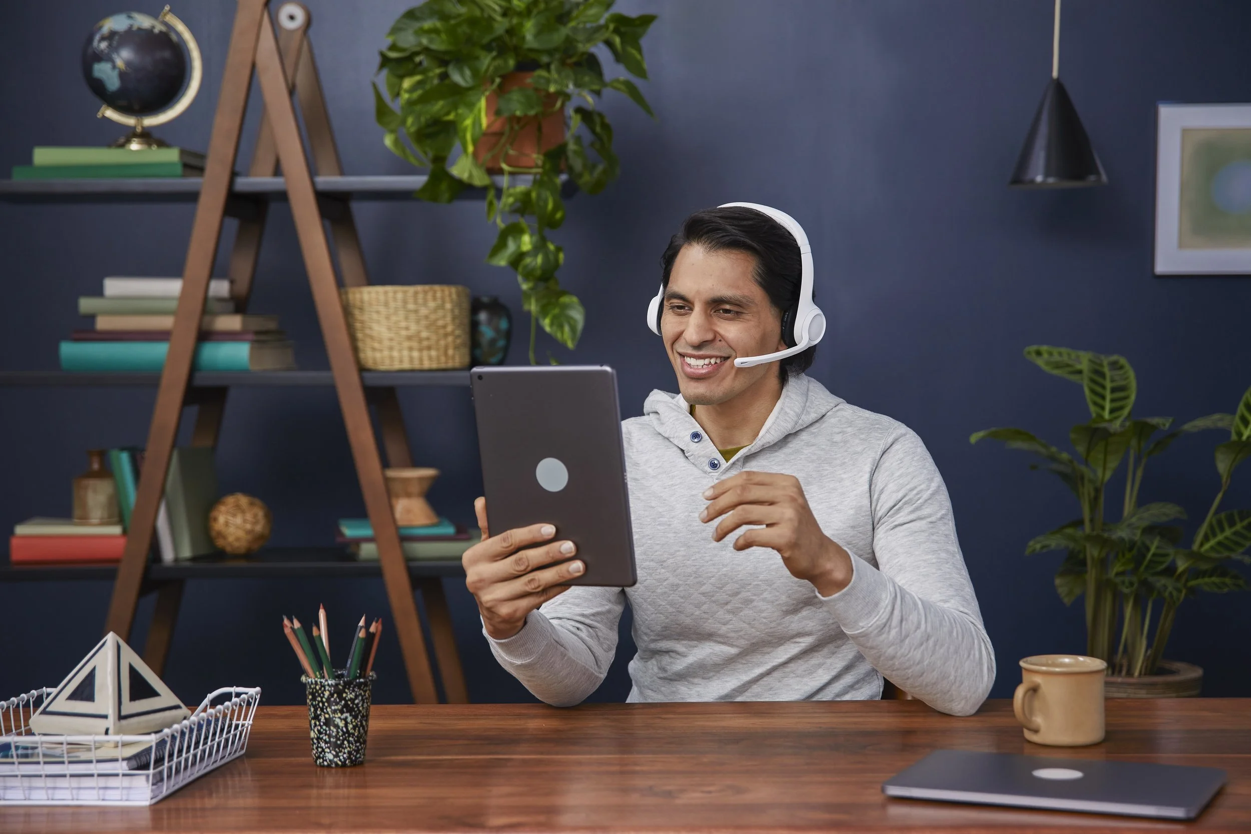 A man in a gray hoodie wearing a white headset smiling while looking at a tablet in his hand, sitting at a wooden desk with decorative and organizational items, surrounded by plants and books in a home office.