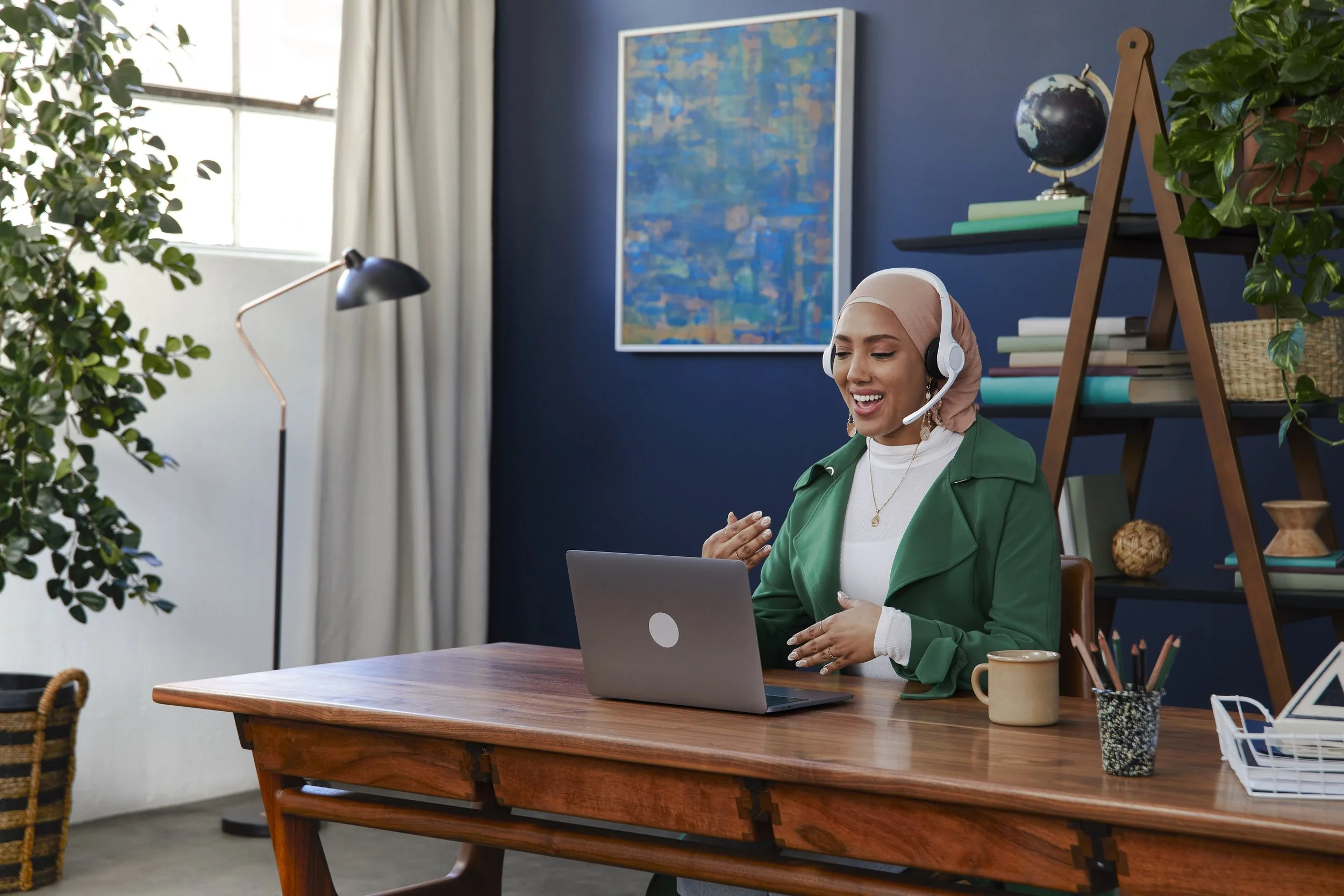 A woman wearing a beige headscarf and green blazer, with headphones, smiling while sitting at a wooden desk with a laptop in a bright room with a blue wall, bookshelf, and large window.
