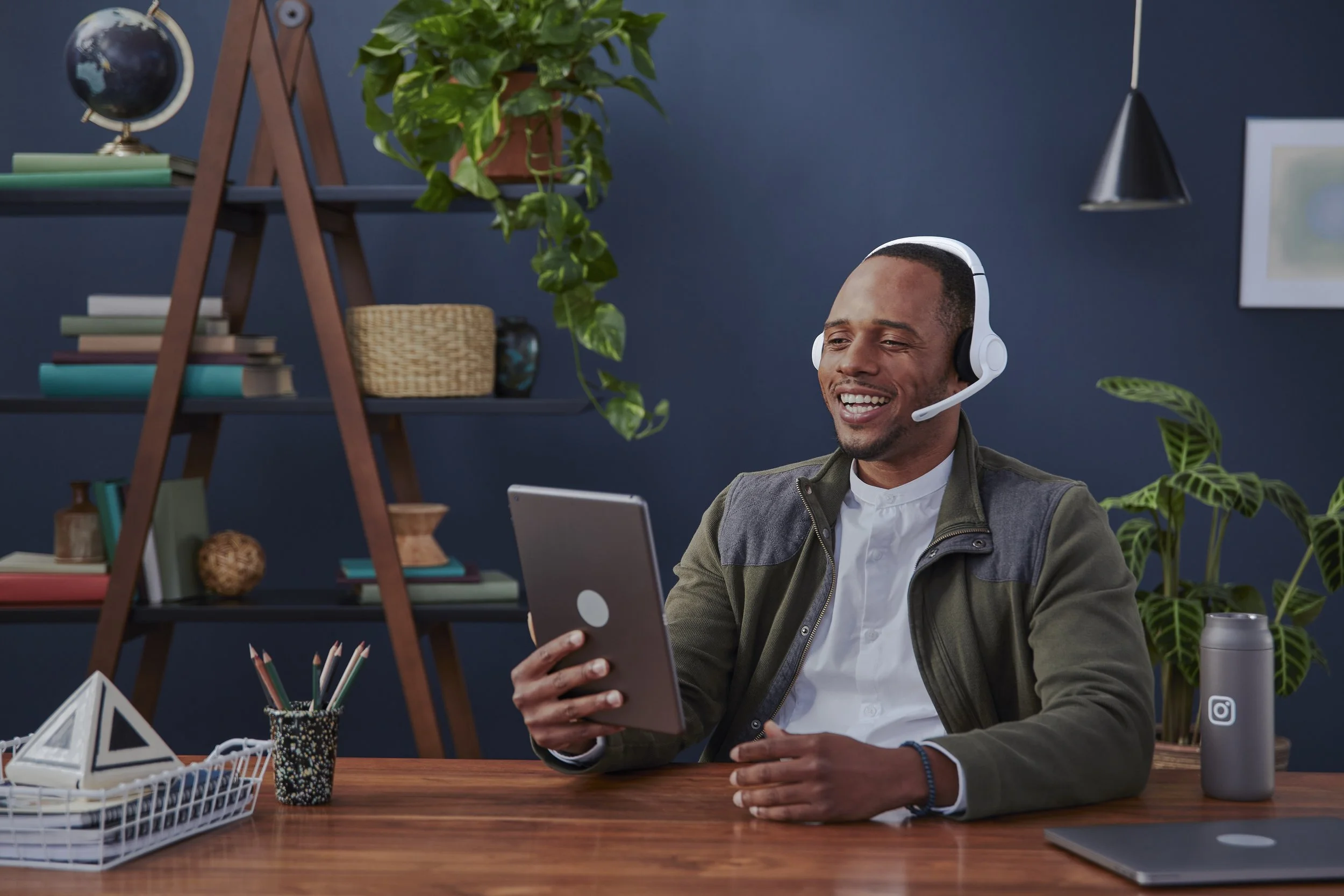 A man sits at a desk, wearing a white headset, smiling while looking at his tablet. The desk has a basket of colored pencils, a cup, and a water bottle. Behind him, there are books, a globe, plants, and a blue wall.