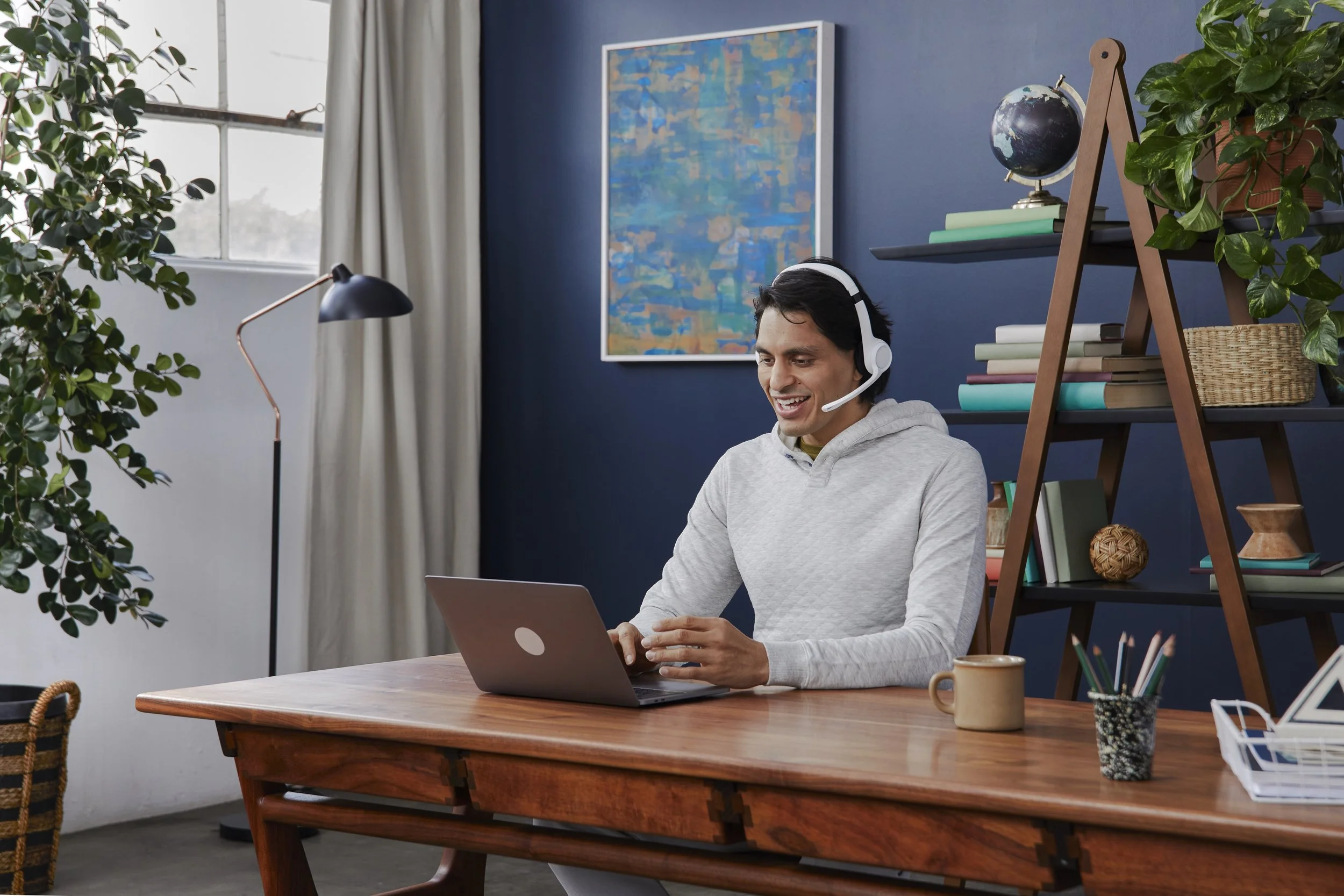 A man sitting at a wooden desk working on a laptop, wearing a headset, in a room with a blue wall, a large window with curtains, green plants, and a black shelf with books and decorative items.