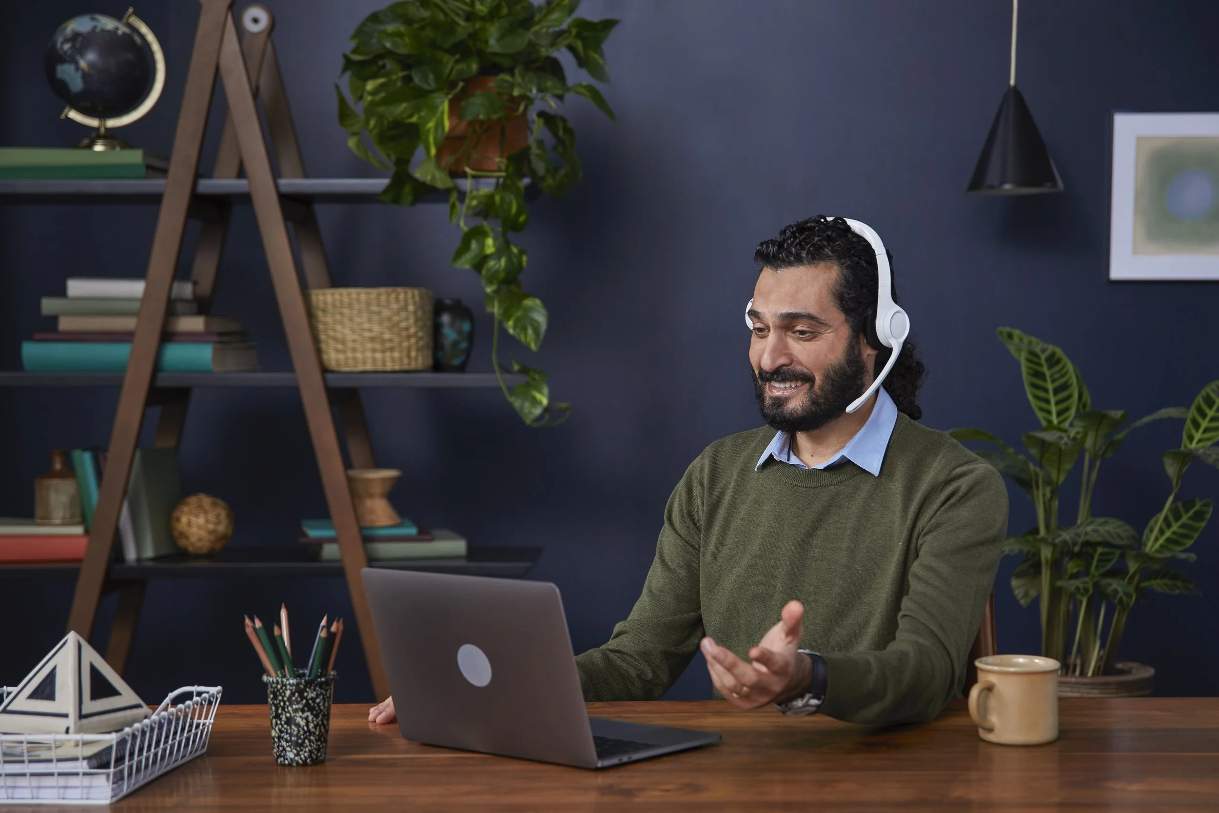 A man with dark, curly hair, a beard, and a mustache sitting at a desk with a laptop, wearing a green sweater and a white headset, smiling and gesturing with one hand. The desk has a mug, a container with colored pencils, and a wire basket with paper