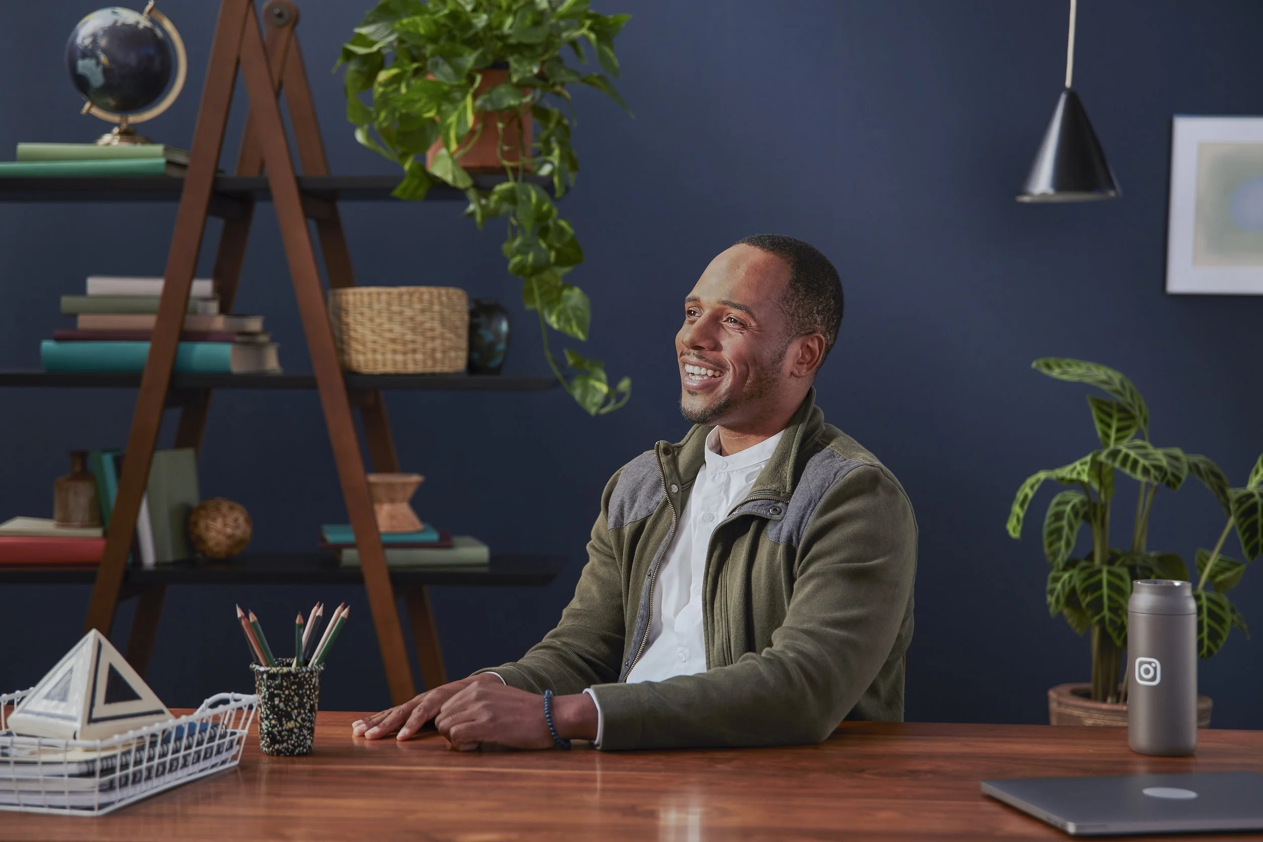 A man with short dark hair and a beard, smiling, sitting at a desk in a modern office or home workspace with a blue wall, surrounded by green plants, books, and desk accessories.