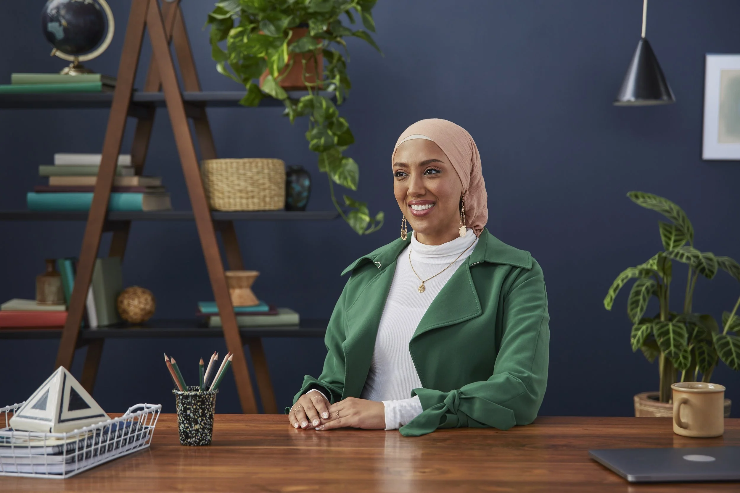 A woman wearing a pink headscarf and green jacket sitting at a wooden desk with plants and office supplies in a modern home office.