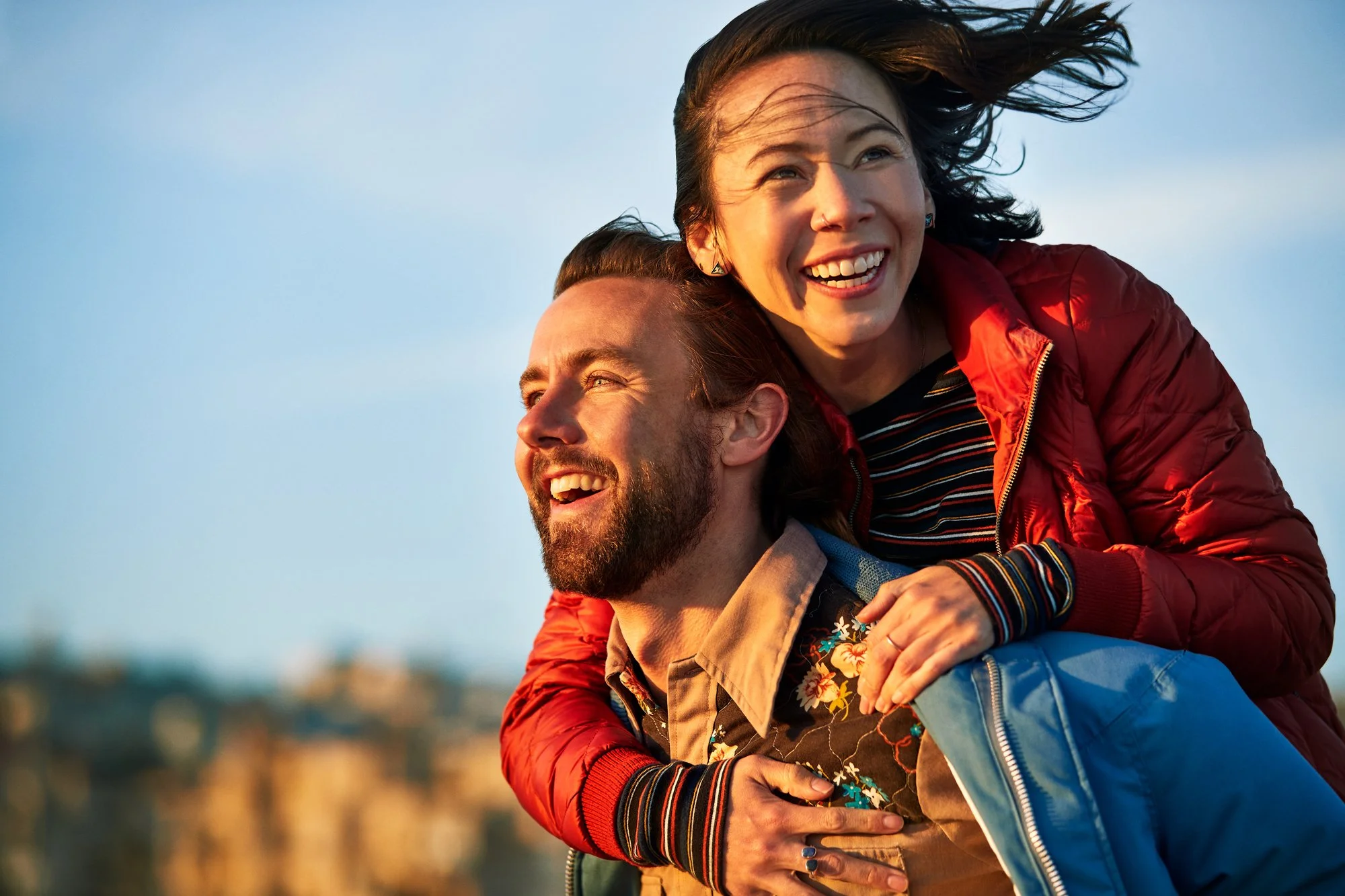 A woman with long hair happily riding piggyback on a man with a beard in a scenic outdoor setting during sunset.