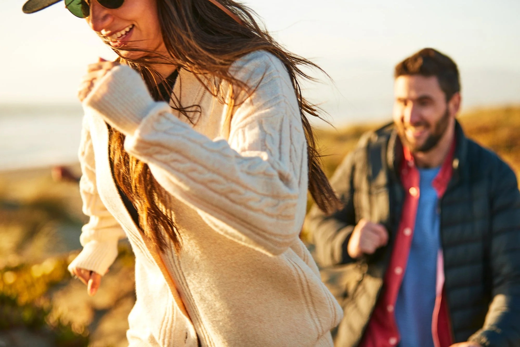A woman with long brown hair wearing sunglasses and a cream-colored sweater, smiling and enjoying herself outdoors during sunset, with a man in the background smiling and in a dark jacket.