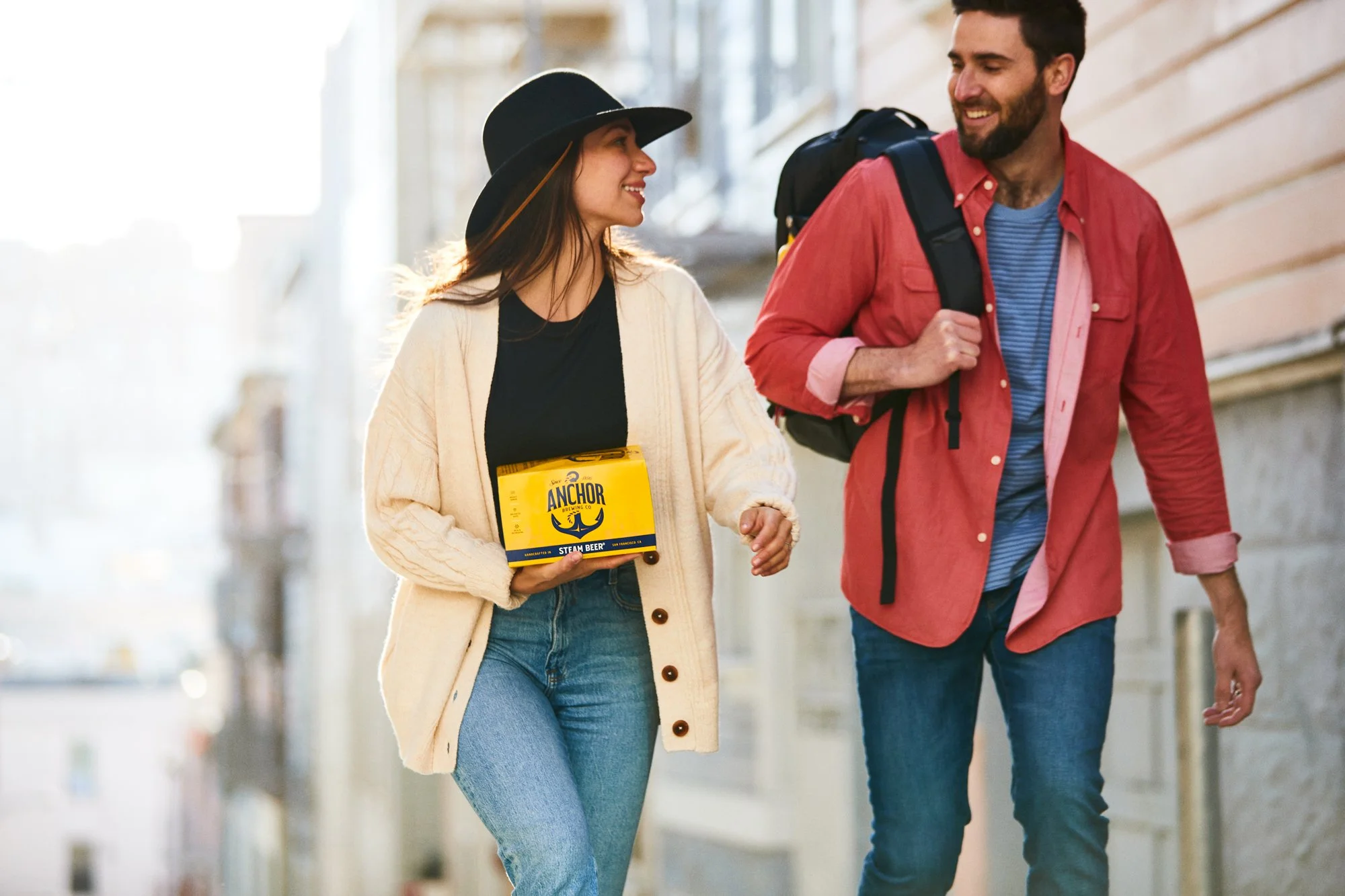 A young woman and man walking and talking on a sunny day. The woman is wearing a black hat, cream-colored cardigan, black shirt, and jeans, holding a pack of Anchor Brewing Company Steam Beer. The man is wearing a red shirt over a blue t-shirt, carry