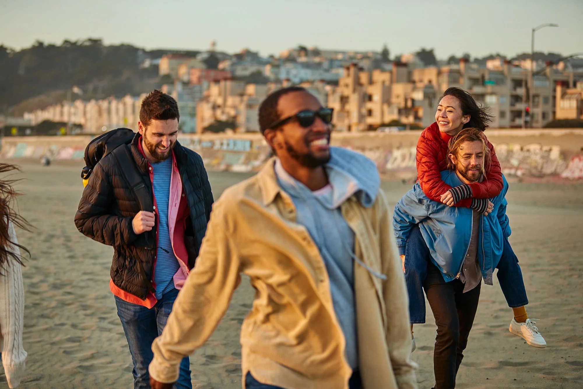 A group of four diverse friends walking along the beach during sunset, smiling and enjoying each other's company, with a residential area visible in the background.