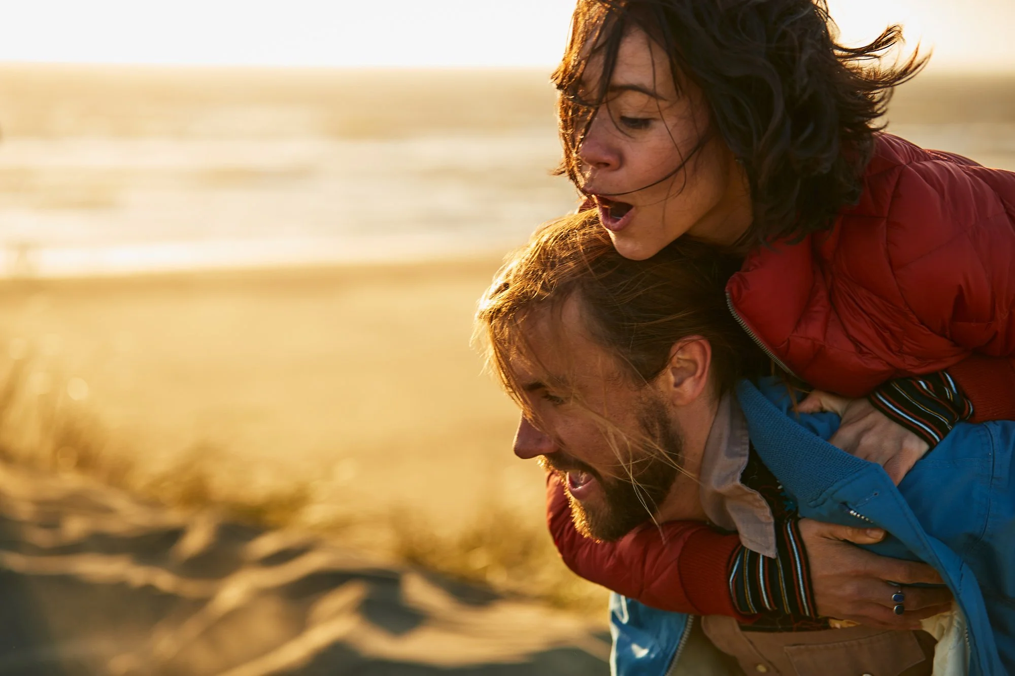 A woman with dark curly hair in a red jacket is riding on the shoulders of a man with blond hair in a blue jacket at the beach during sunset. The woman looks surprised or excited.