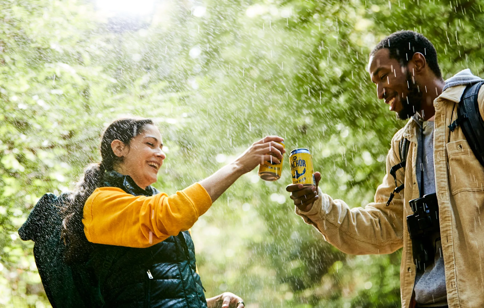Two people, a woman and a man, sharing a toast with cans of beer in a rain shower in a forest. The woman wears an orange jacket and a black vest, smiling, and the man wears a tan jacket with a backpack, smiling as well. Rain is falling around them an