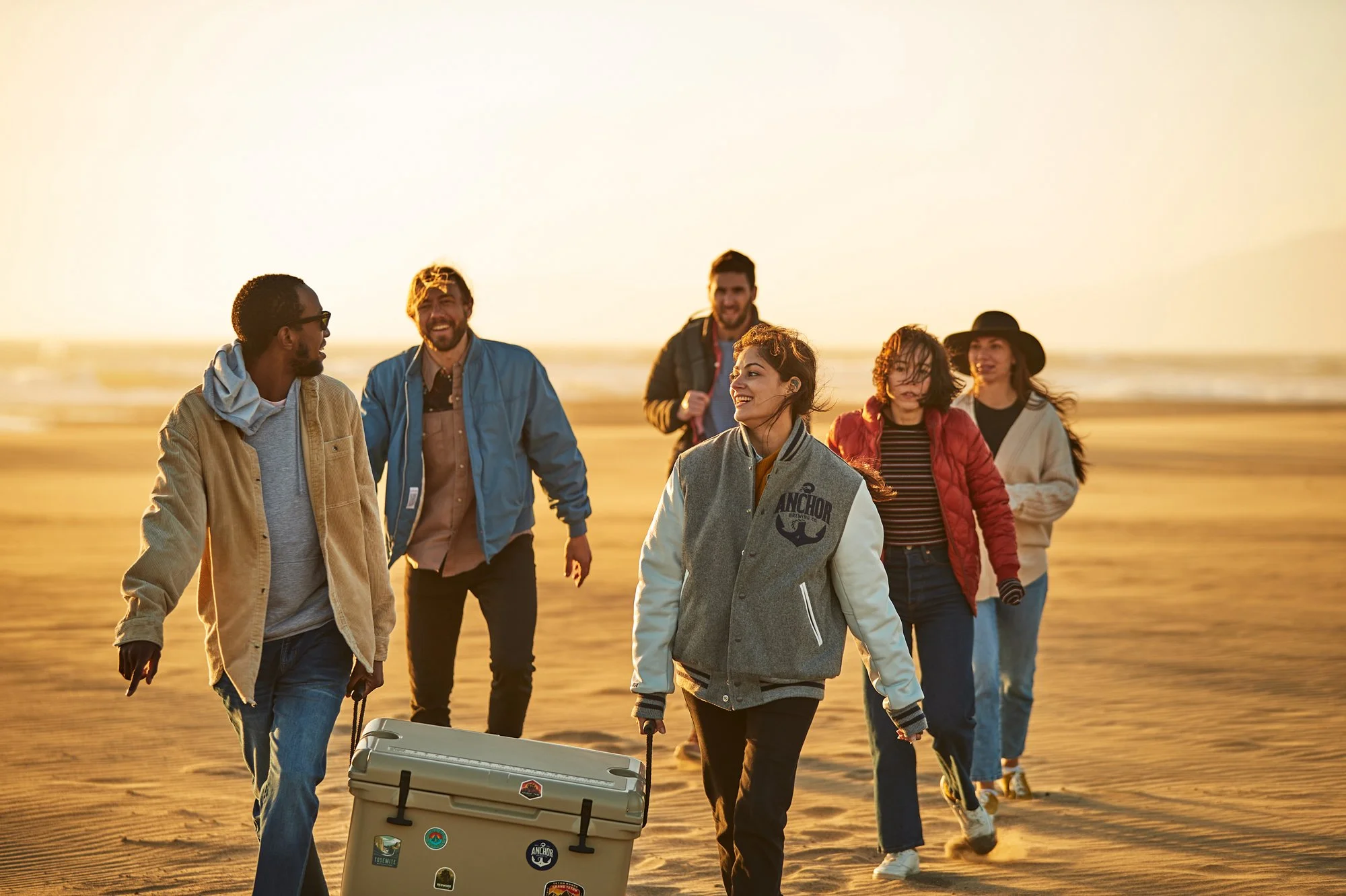 A group of six diverse friends walking on a beach at sunset, pulling a large cooler behind them, and smiling as they enjoy a day by the water.
