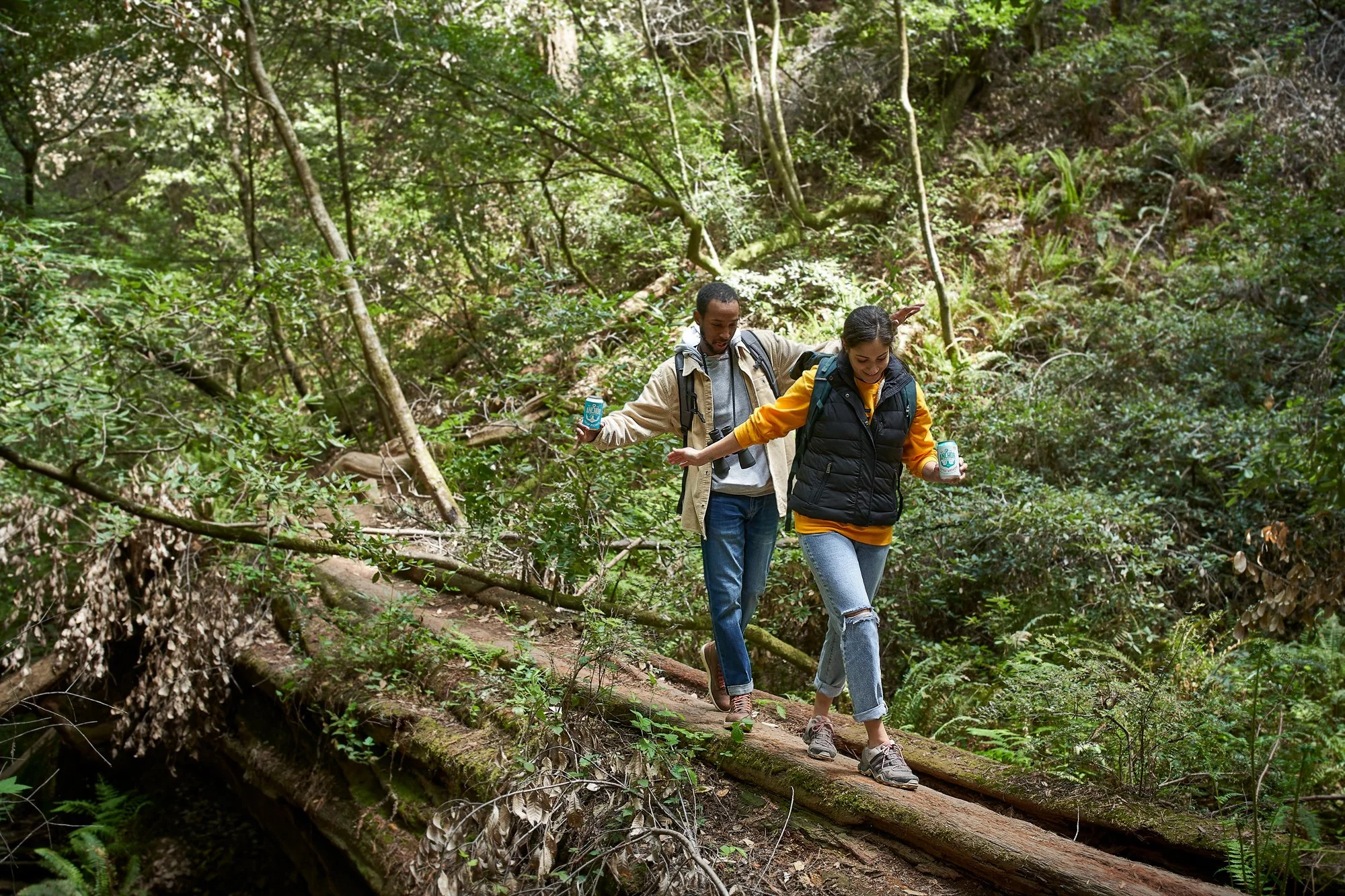 Two people hiking on a trail covered with fallen logs in a lush green forest. They are balancing on a large log across the trail, holding onto cans of Anchor beer, with backpacks and casual clothing.
