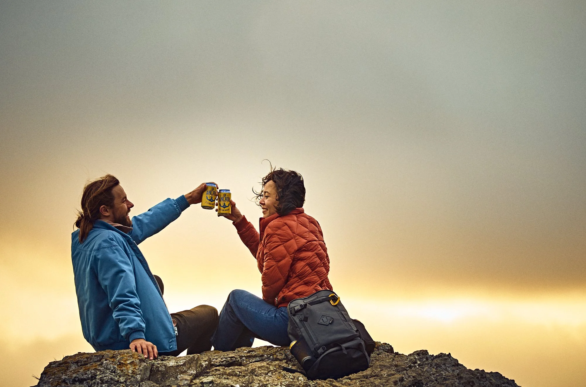 A man in a blue jacket and a woman in a red jacket sit on a large rock outdoors during sunset. They are smiling and holding cans of Anchor beer in a toast. The woman is sitting with her legs crossed on the man's knee, and there's a backpack and a rol