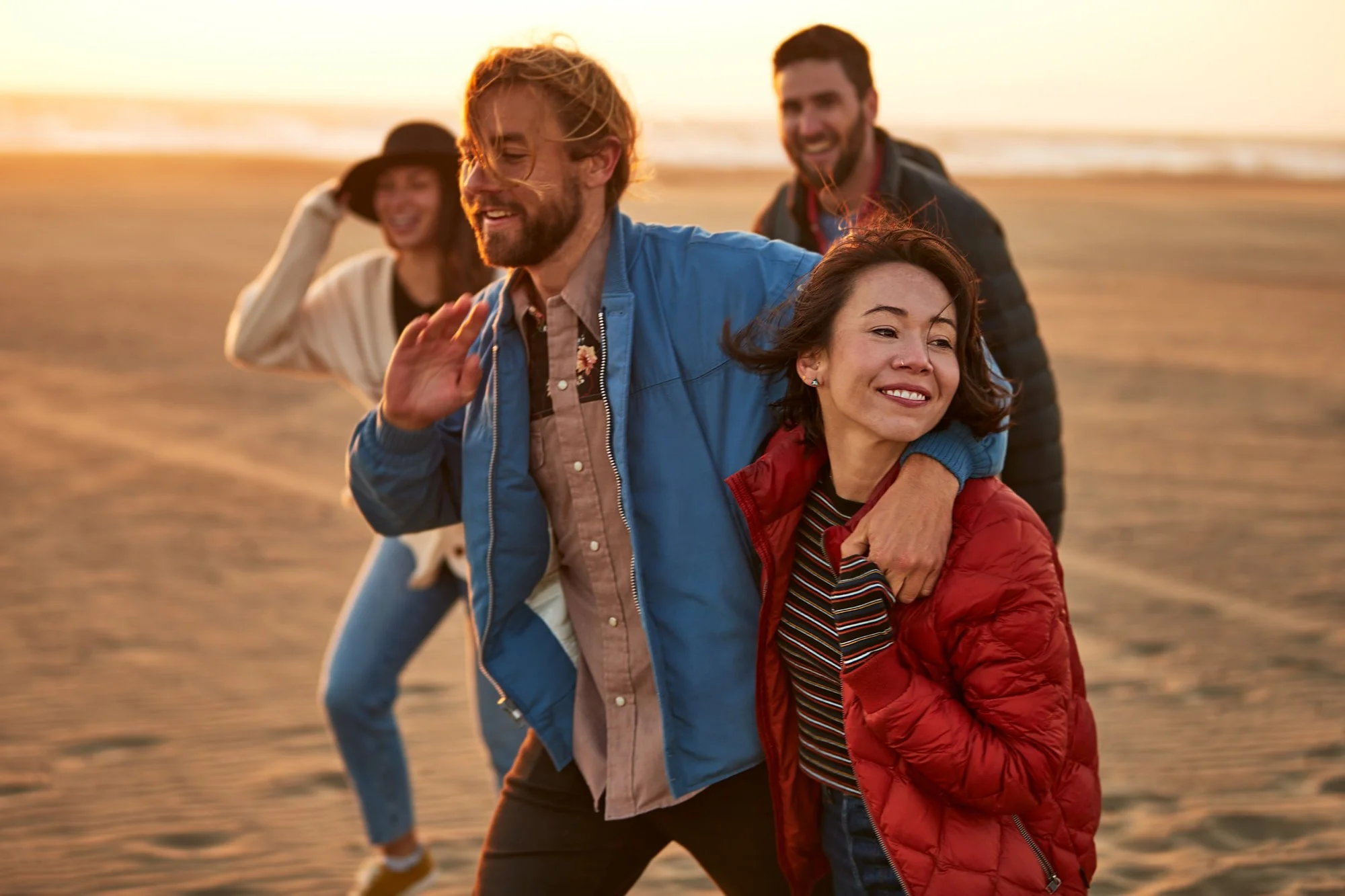 Four friends laughing and playing on a beach during sunset, with the man in blue leading and the woman in red smiling, others following behind.