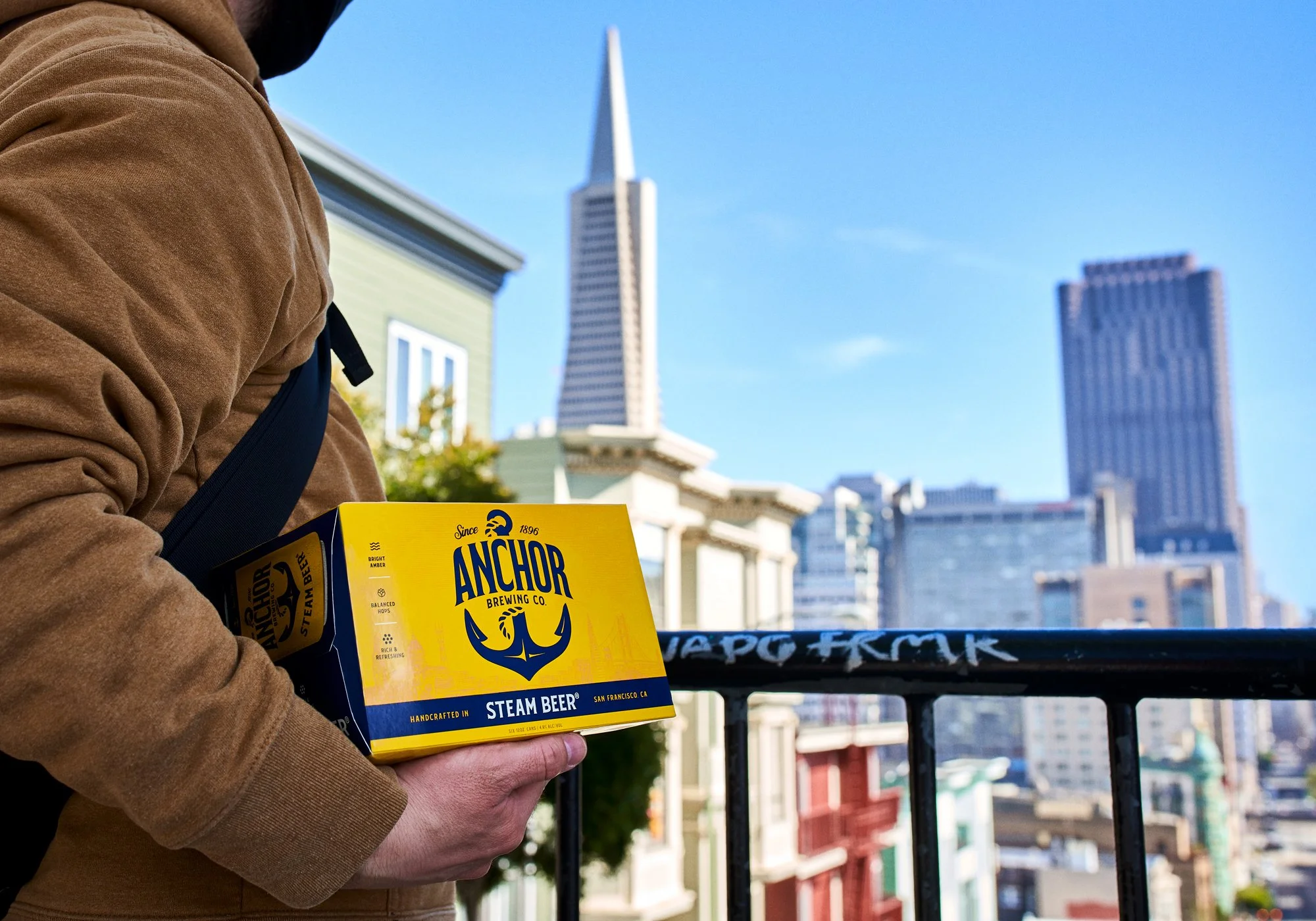 Person holding a yellow box of Anchor Brewing Co. Steam Beer with San Francisco skyline in the background.