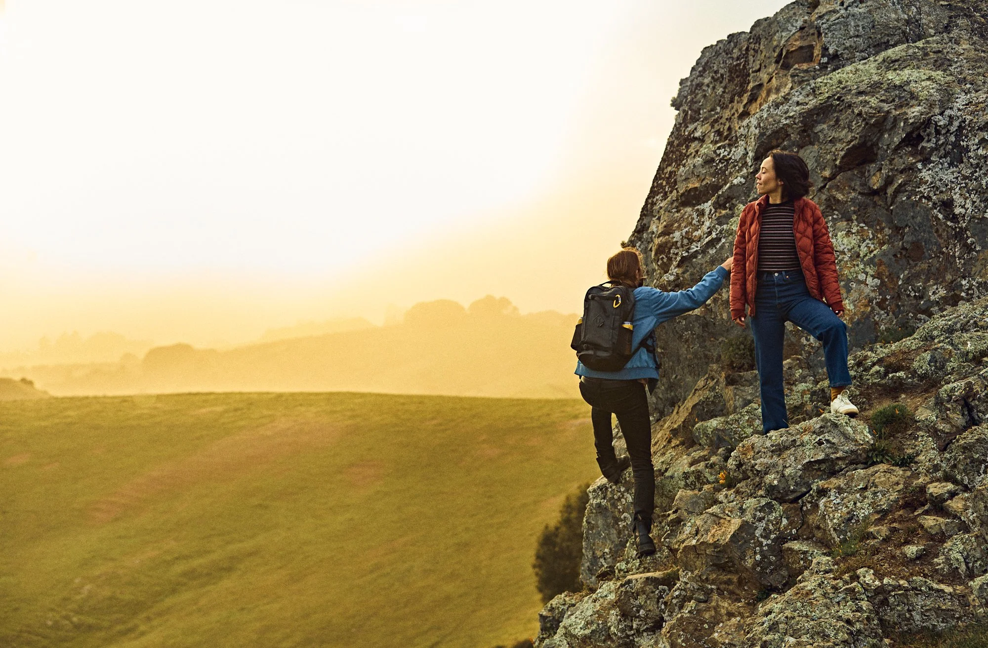 Two women are climbing a rocky outdoor incline during sunset. One woman is helping the other by holding her hand as she ascends. The background features open fields and a soft, golden sky.