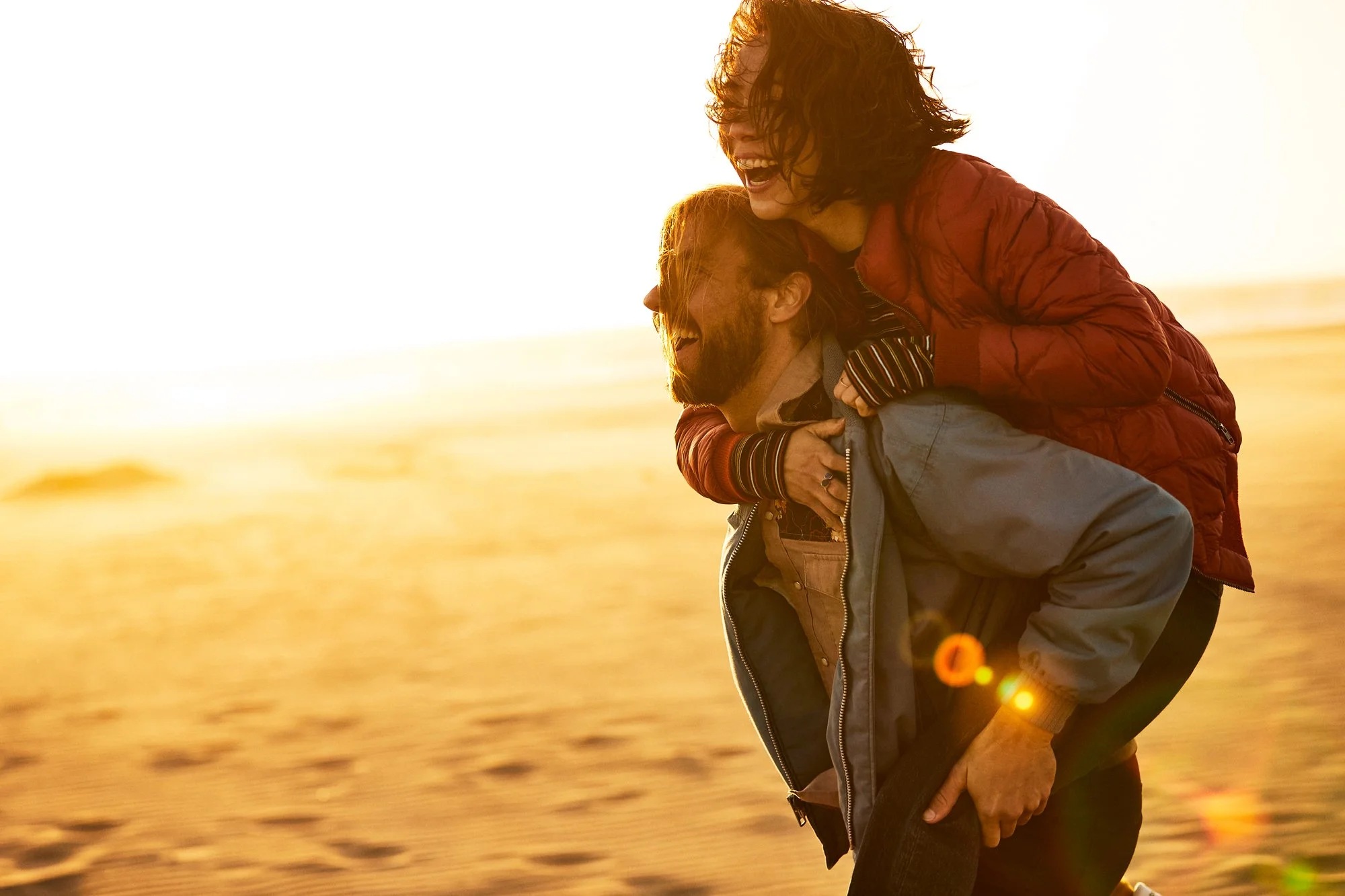 Two people, a man and a woman, enjoying a joyful moment at the beach during sunset, with the woman riding on the man's back.