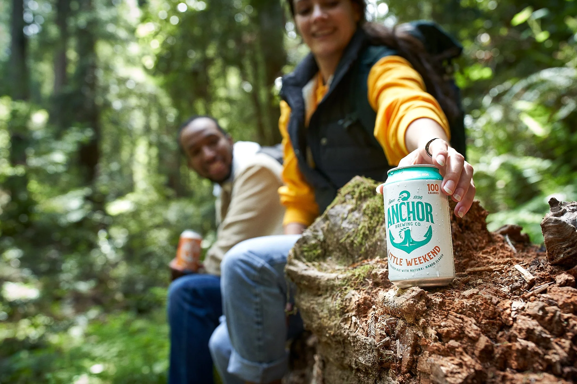 Two people sitting on a fallen tree in a forest, smiling, with a can of Anchor Brewing Little Weekend mango-flavored hard seltzer in the foreground.