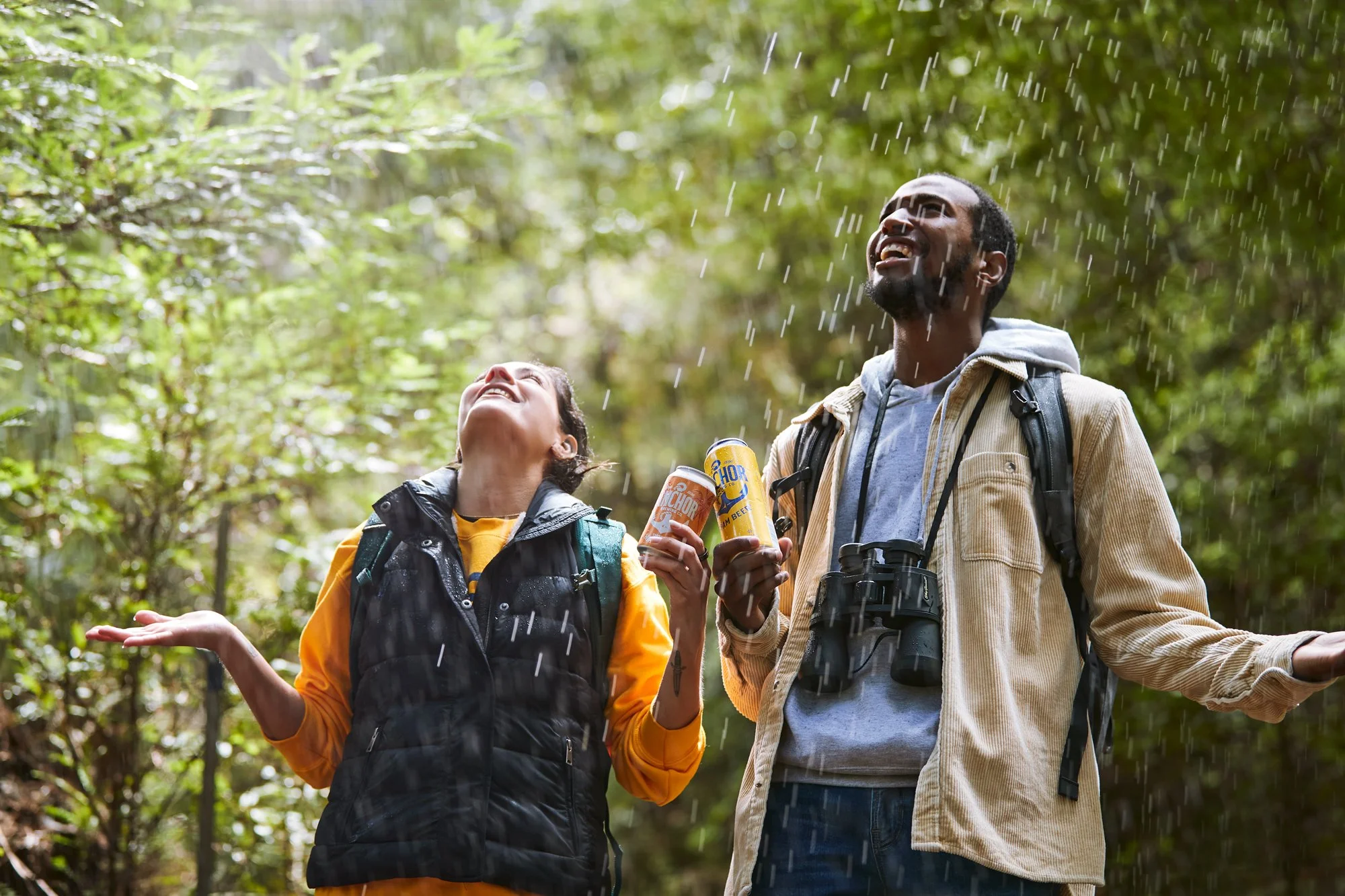 Two hikers, a woman and a man, standing in a forest during rain, smiling and enjoying the moment. The woman wears a yellow shirt and black vest, holding a can of soda. The man has a beige jacket, a gray hoodie, a camera around his neck, and a backpac
