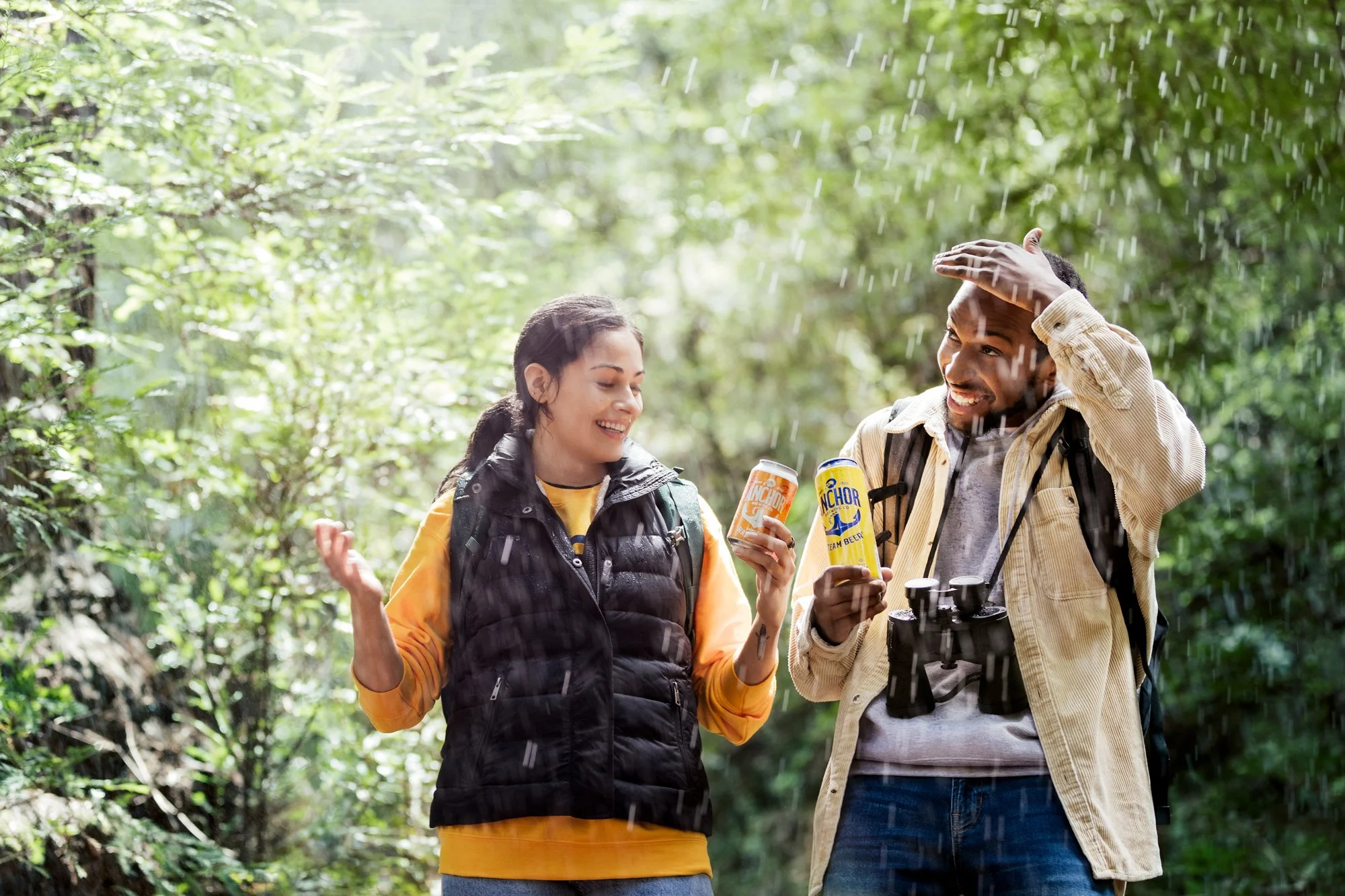 Two hikers, a woman and a man, enjoy a moment in the woods, holding cans of beer, smiling, and chatting. The woman wears a black vest over an orange shirt, and the man wears a beige jacket with binoculars hanging around his neck. Raindrops fall as th