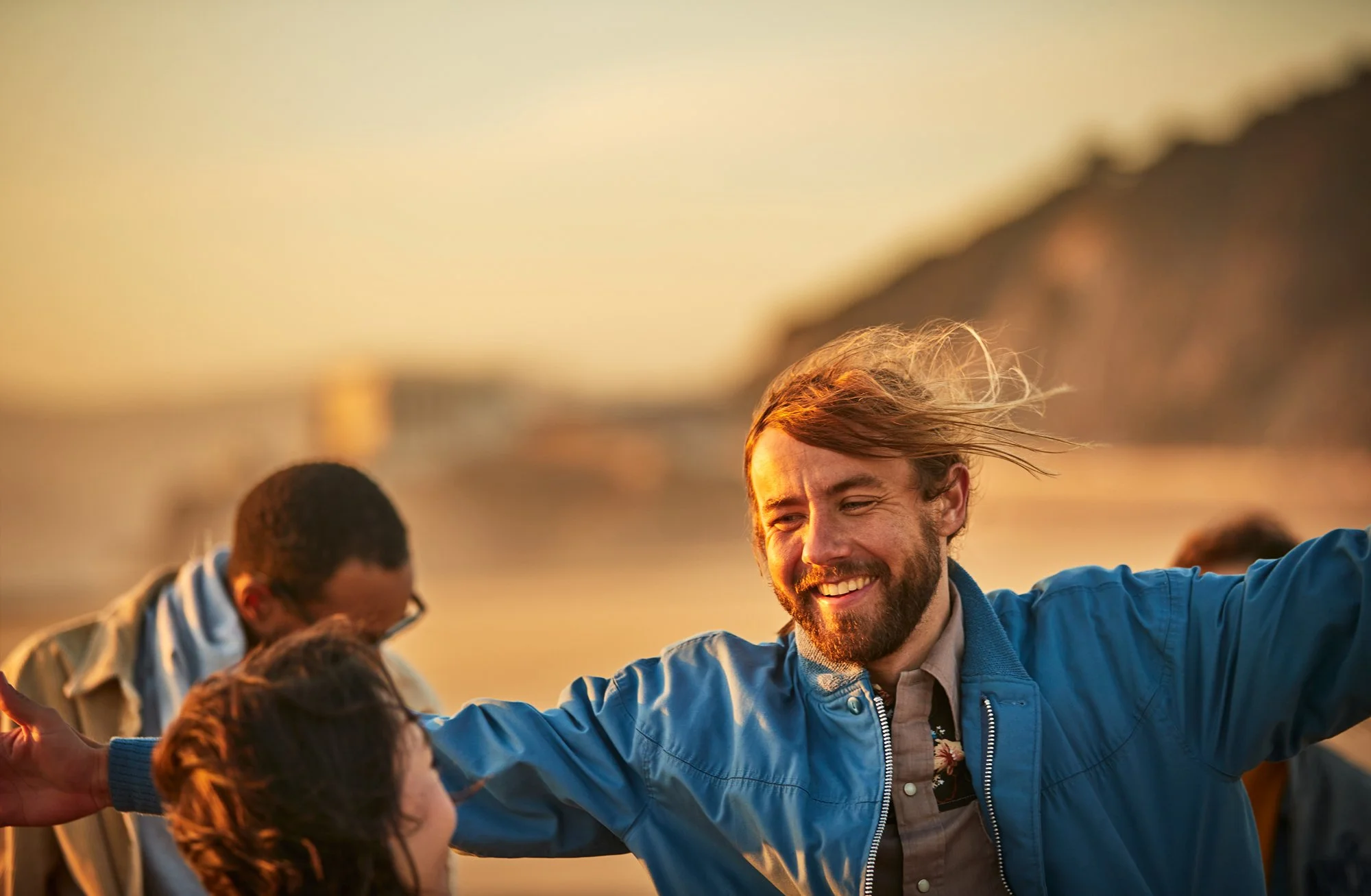 Smiling man with long hair and beard wearing a blue jacket, with a group of people outdoors during sunset, engaging in a cheerful activity.