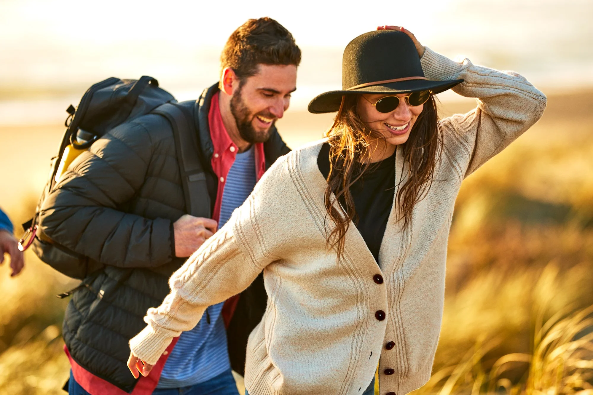 A man and a woman laughing and walking together outdoors during sunset, the woman is wearing a wide-brimmed hat and sunglasses, and the man has a backpack.