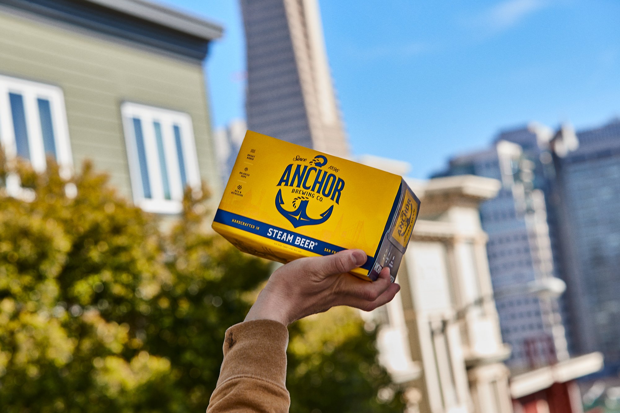 A person holding a yellow box of Anchor Brewing Co steam beer in an urban outdoor setting with buildings and blue sky in the background.