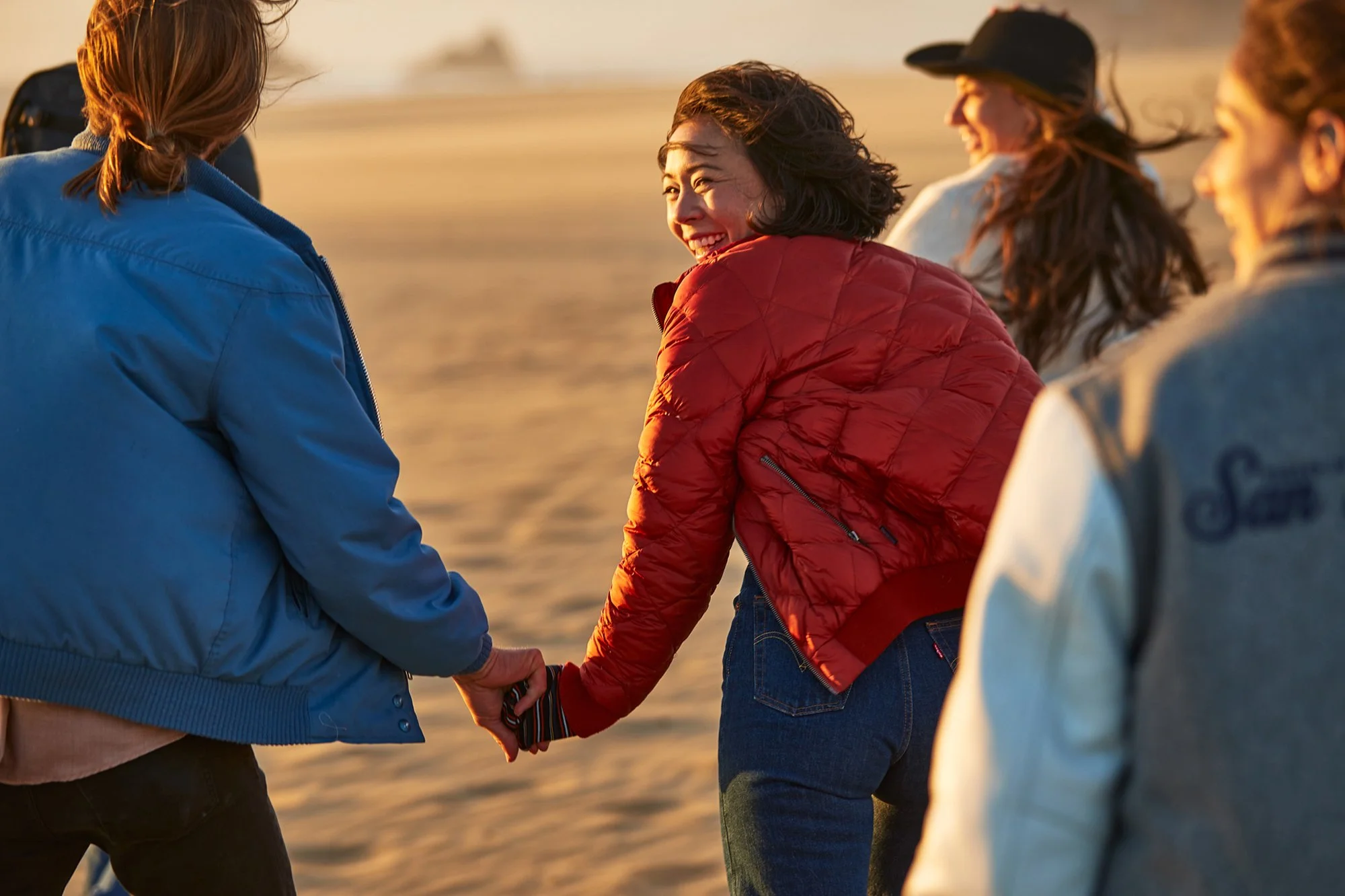 Four women holding hands and smiling on a beach at sunset