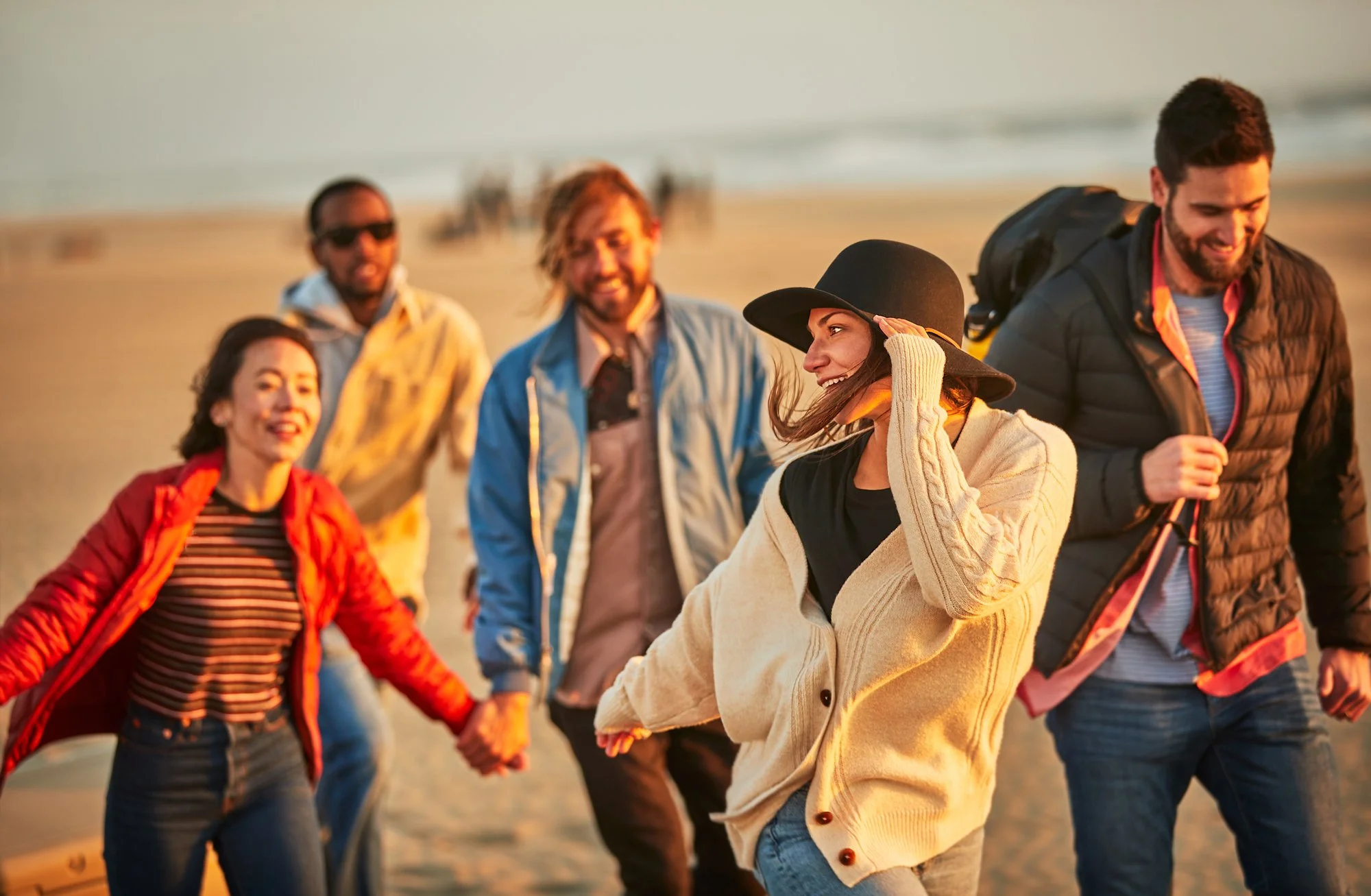 Group of friends holding hands and walking along the beach during sunset, smiling and enjoying each other's company.