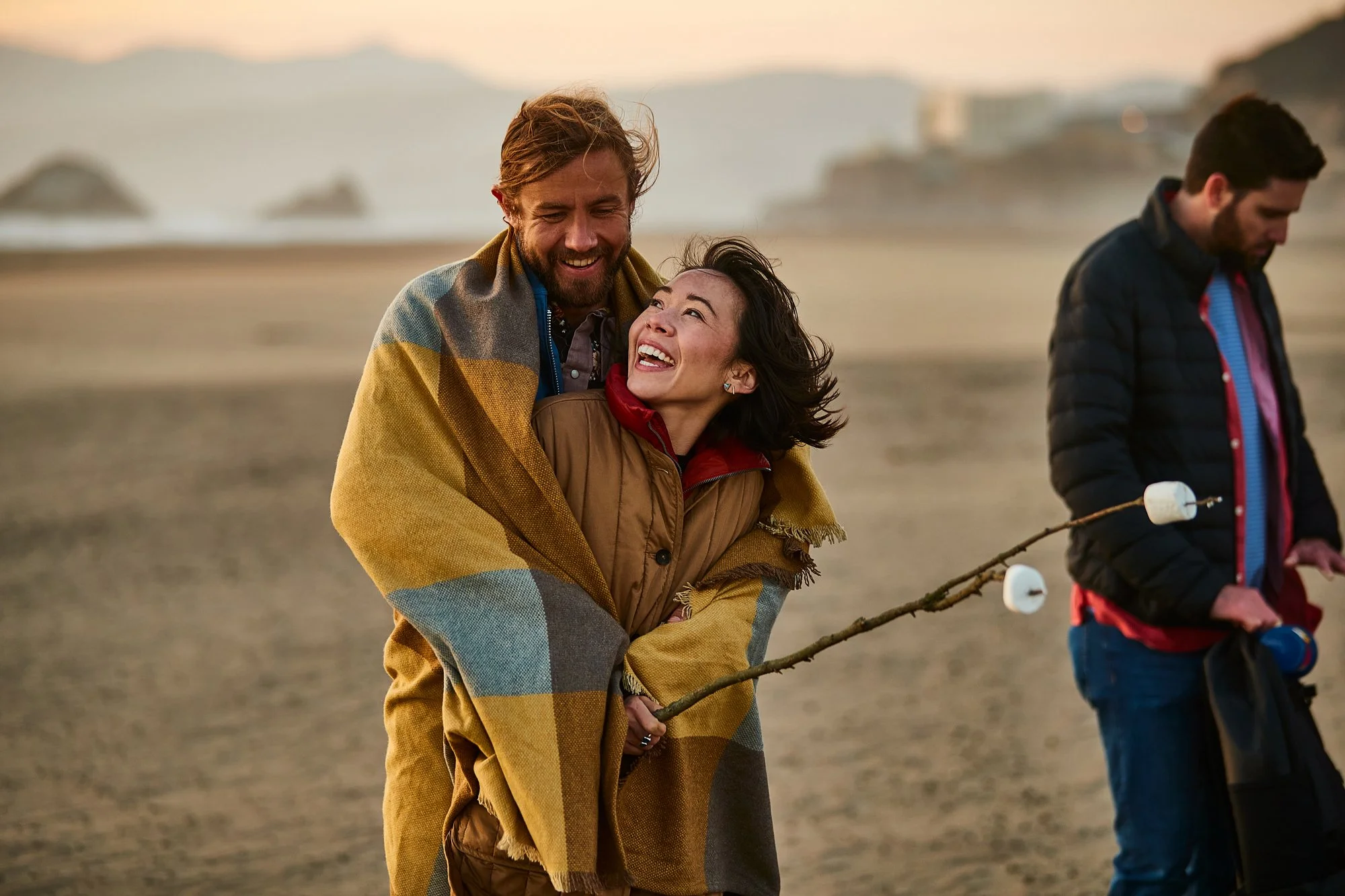 Two smiling people walking on the beach during sunset, wrapped in a colorful blanket, with a man and woman looking happily at each other and another man holding marshmallow sticks nearby.