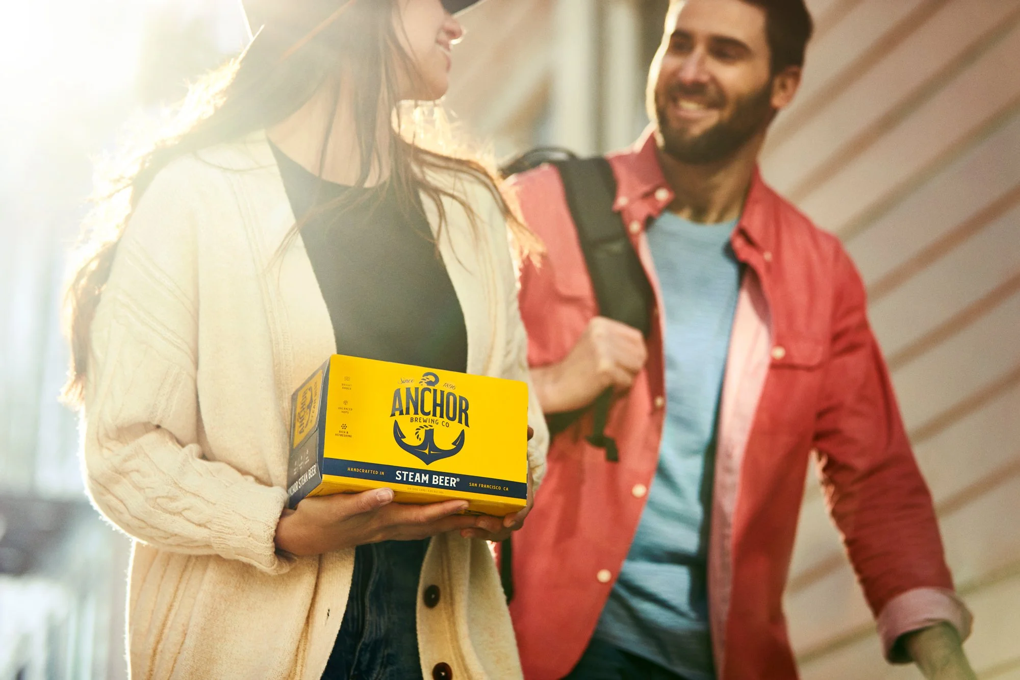 A woman holding a yellow box of Anchor Brewing steam beer, standing next to a man in a pink jacket, both smiling and talking in a well-lit indoor space.